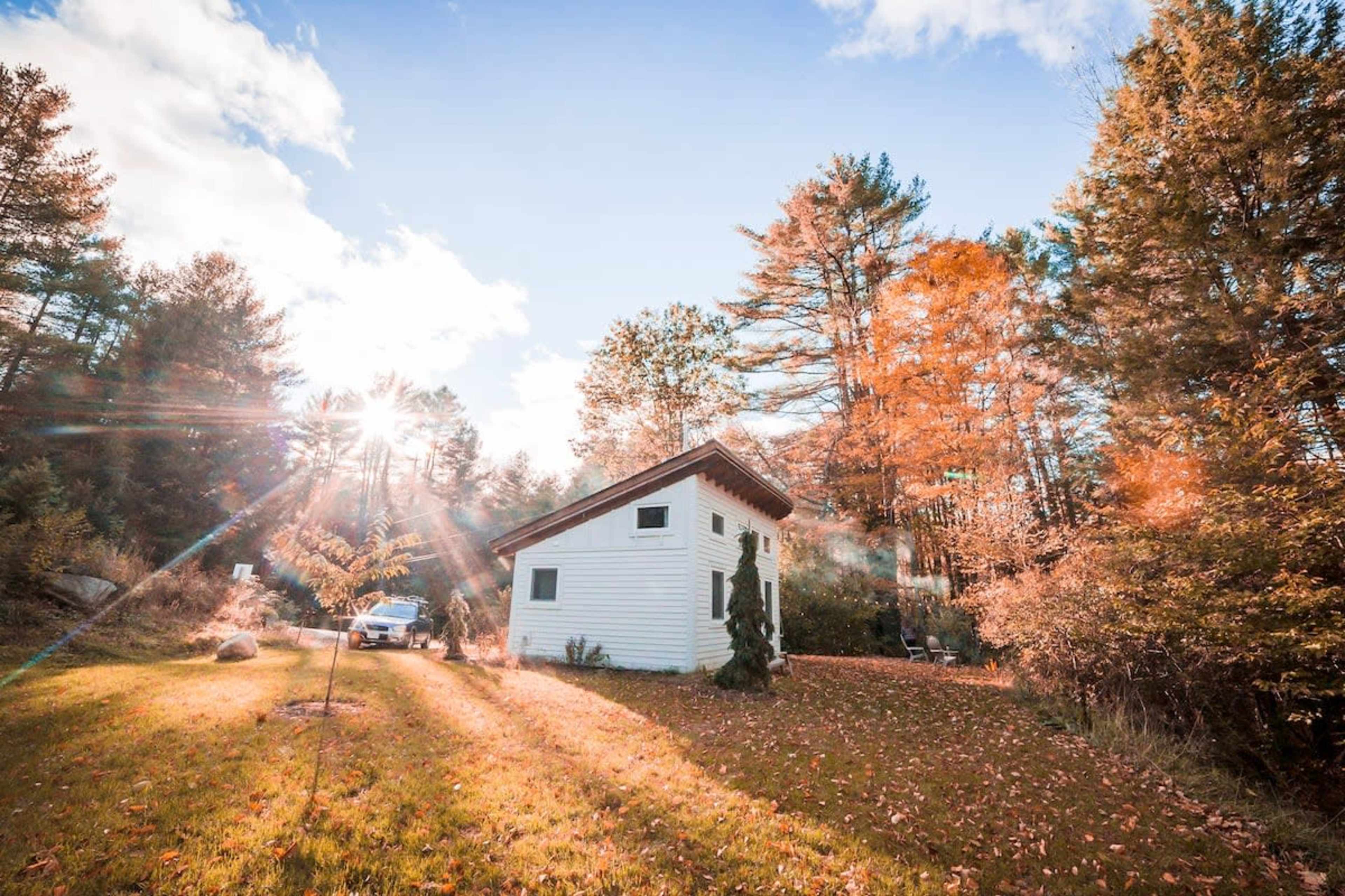 A small white house stands on a grassy area surrounded by trees with autumn leaves, under a bright sky with sunlight streaming through.
