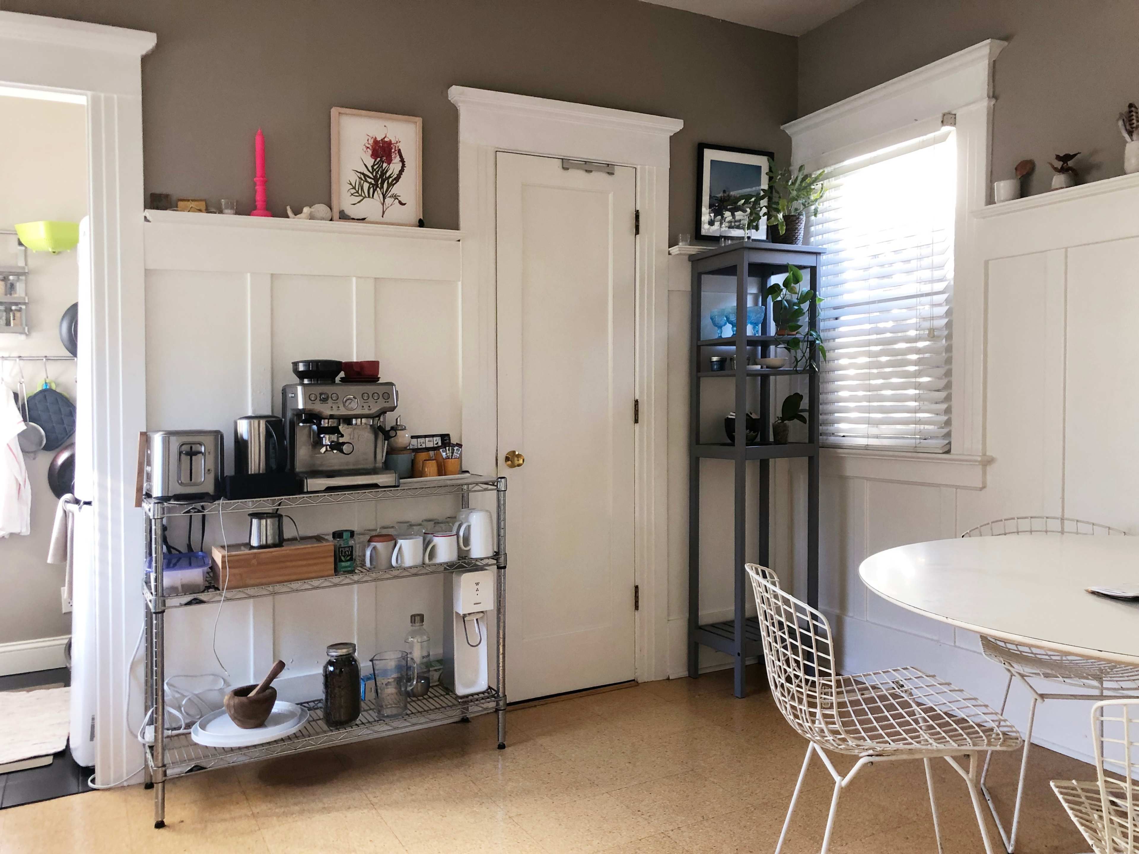 A kitchen area featuring a coffee station, a table with white chairs, and shelves holding plants and decorative items.