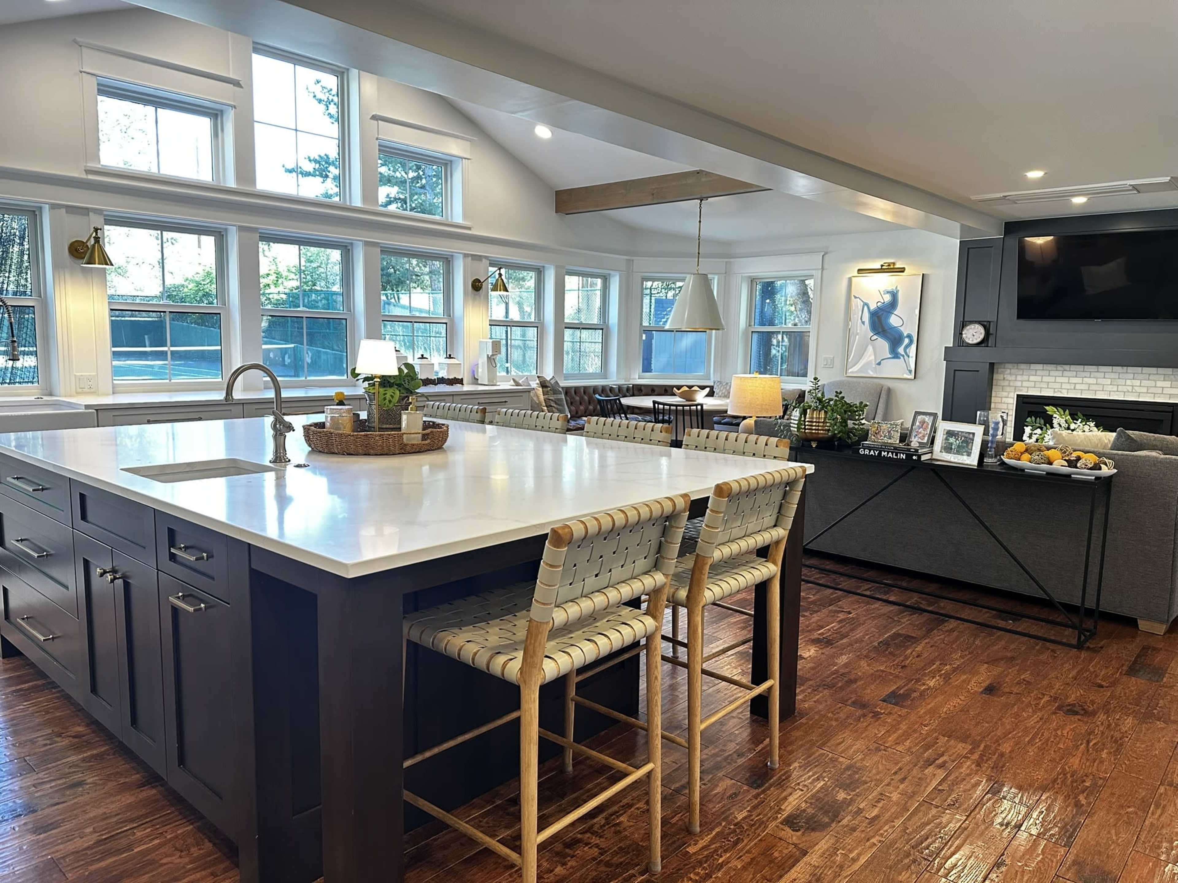 A modern kitchen features a large central island with bar stools, surrounded by natural light from multiple windows, and a cozy seating area with a sectional couch.