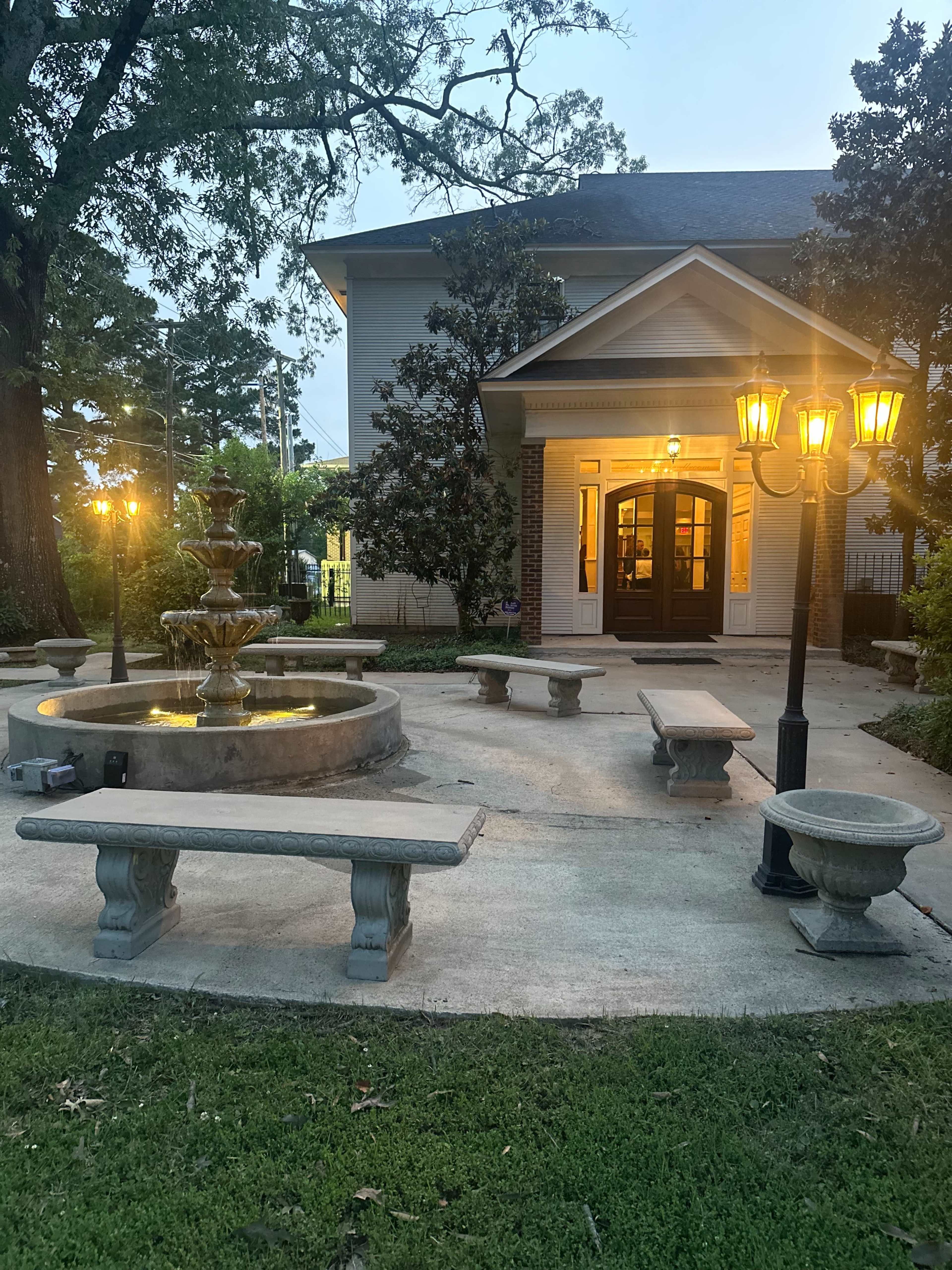 The image shows a courtyard with a fountain, stone benches, and street lamps in front of a building.