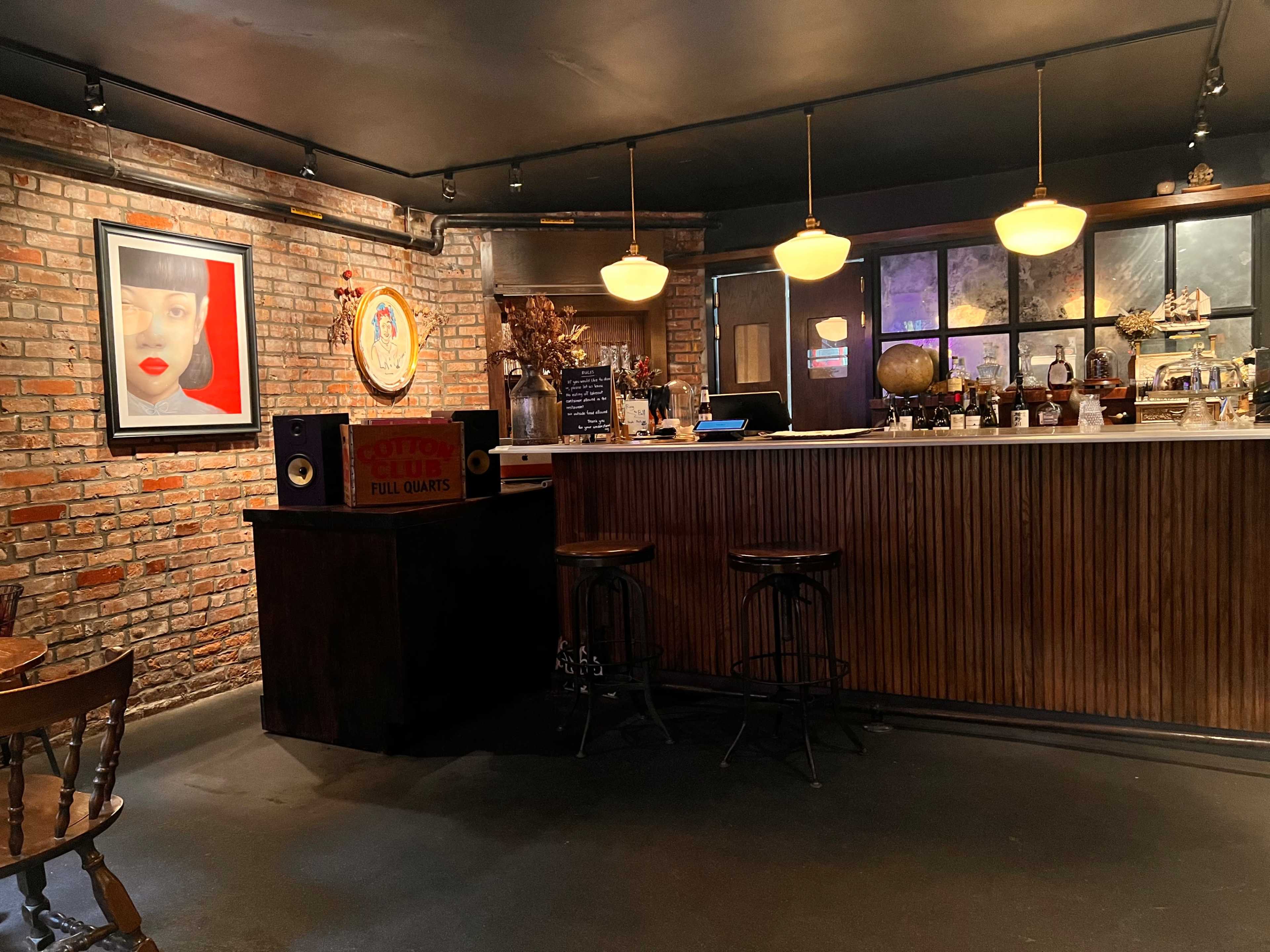 The image shows a rustic bar with a brick wall, wooden counter, and vintage decor, including paintings and a globe.