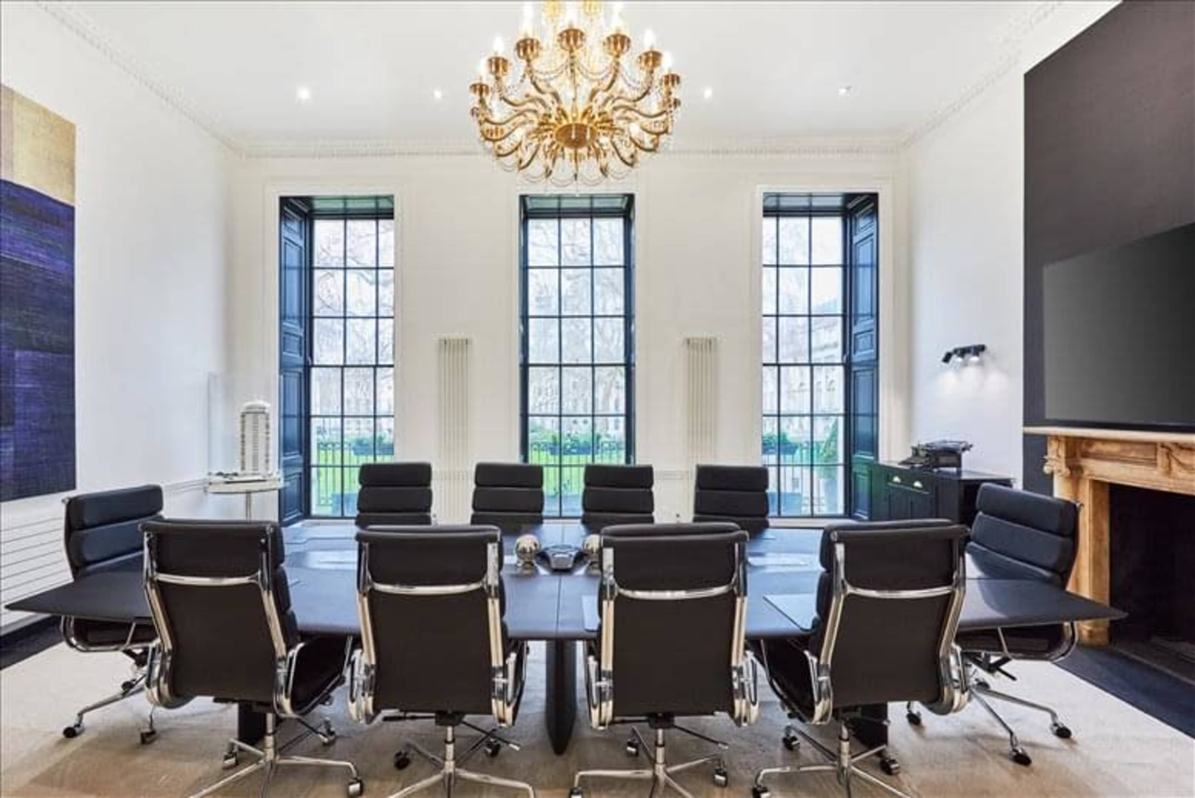 A modern conference room features a long table surrounded by black leather chairs, tall windows allowing natural light, and a large chandelier overhead.