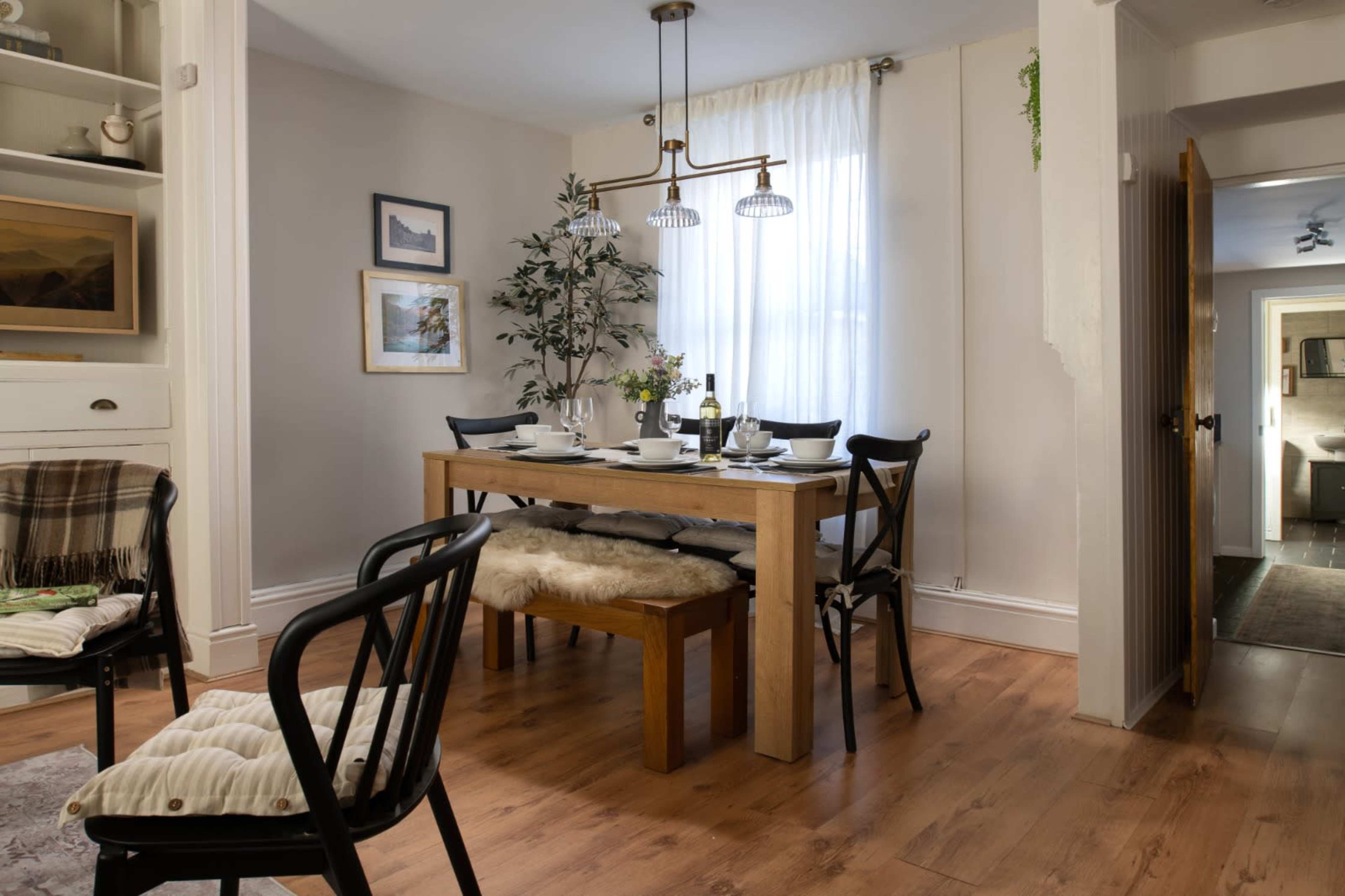 A dining room features a wooden table set for a meal, surrounded by black chairs and a large window with sheer curtains.