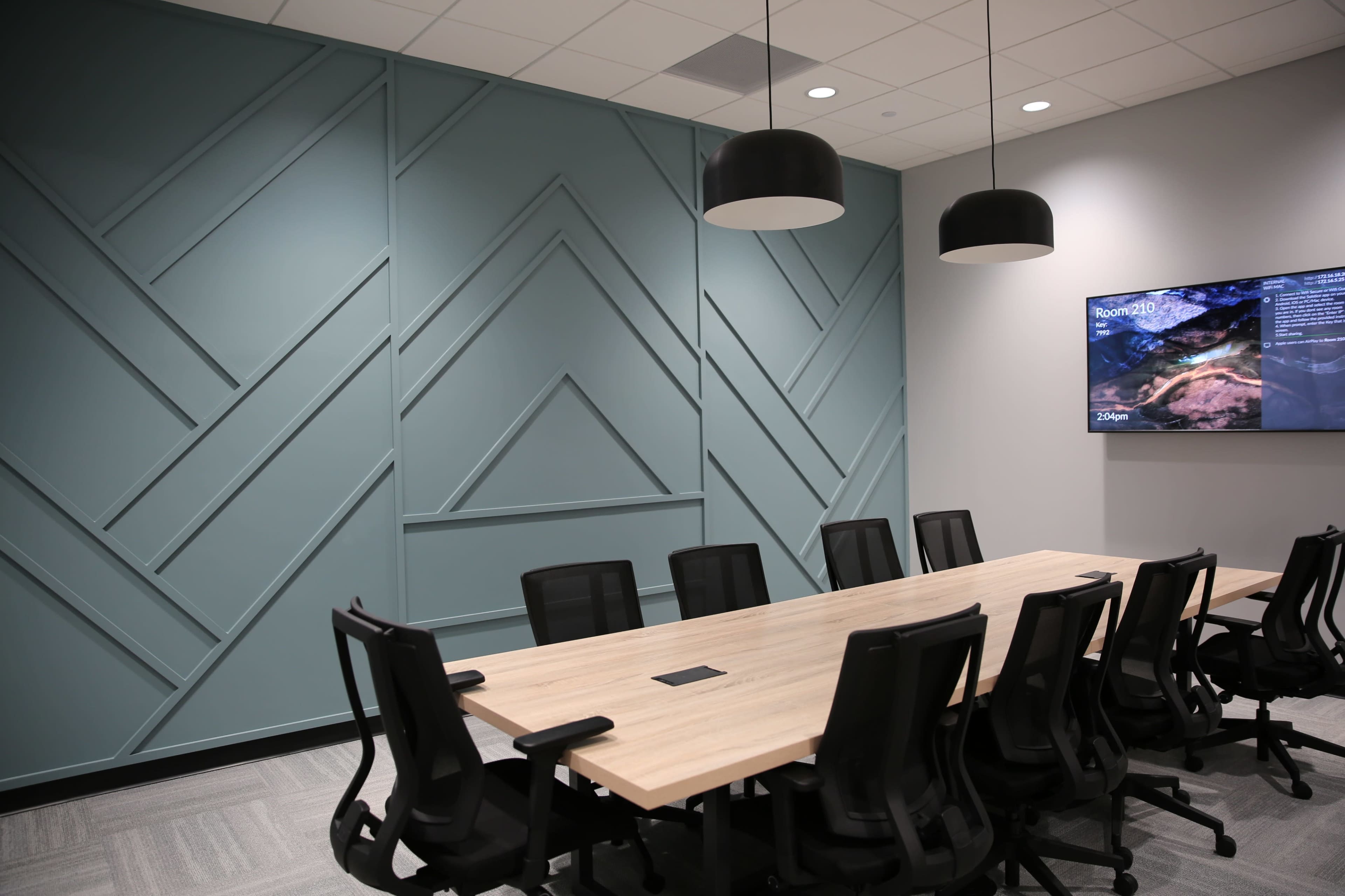 A modern conference room featuring a long wooden table surrounded by black mesh chairs, with a textured blue-green wall and a television screen displaying information on one side.