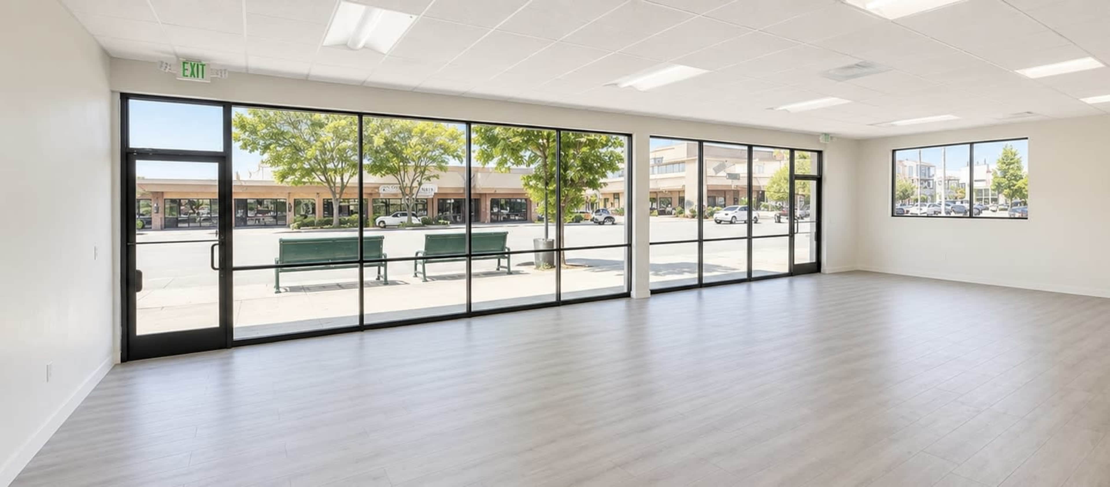 The image shows a spacious, empty room with large glass windows facing a street lined with parked cars and benches.