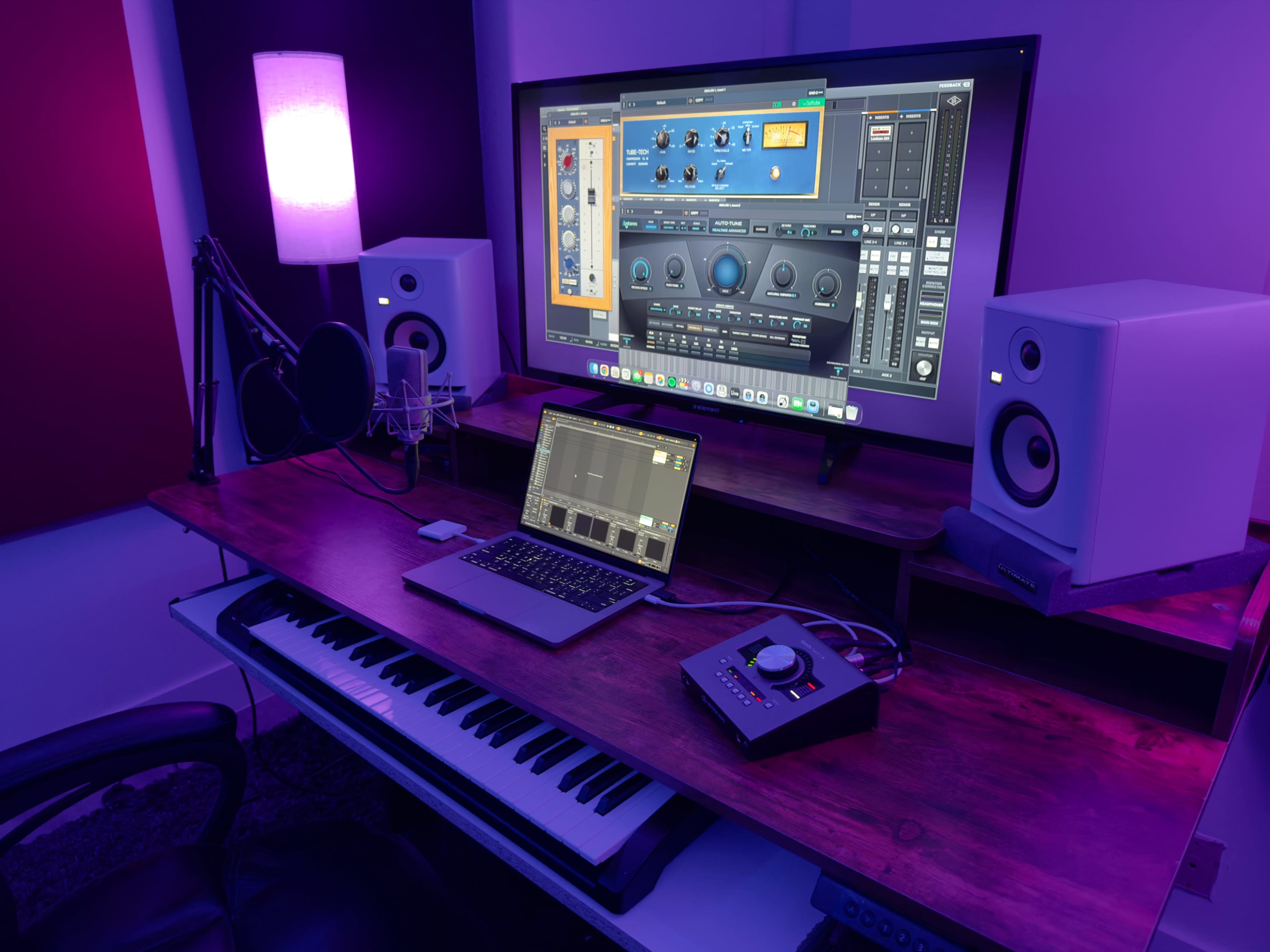 A computer monitor displaying audio software sits on a wooden desk with a keyboard, laptop, and audio interface, flanked by studio monitors and illuminated by purple lighting.
