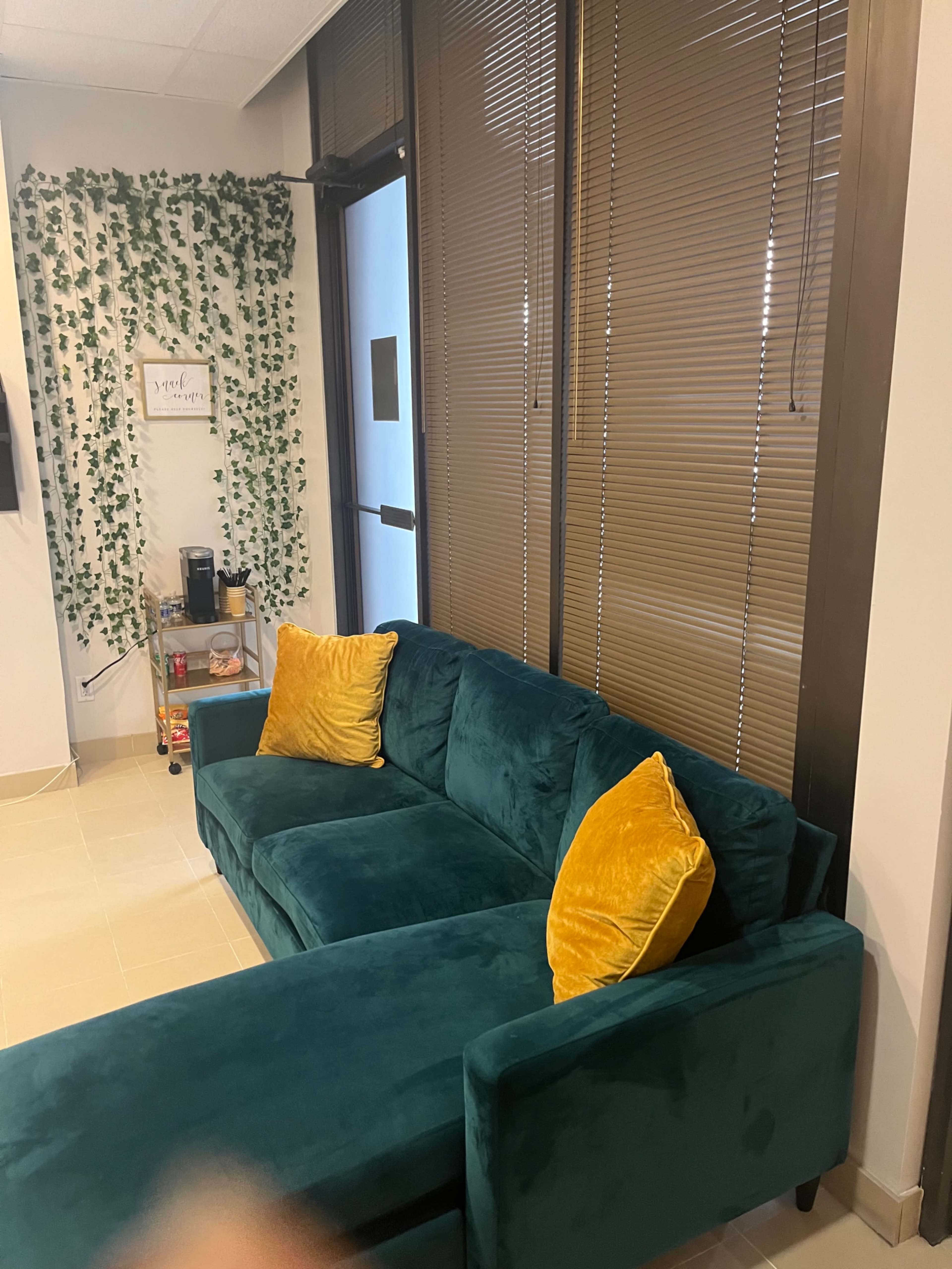 The scene features a teal couch with yellow pillows positioned near a glass door and a wall adorned with green decorative vines.
