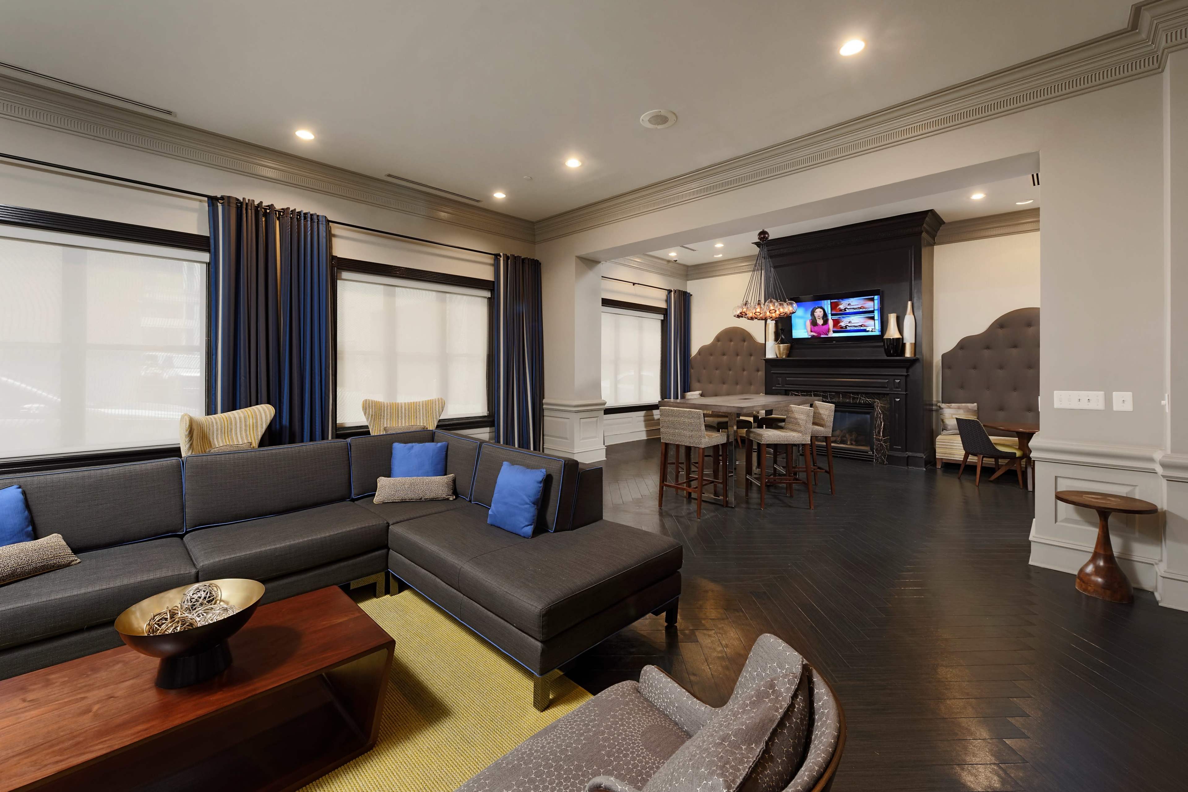 A modern living room featuring a sectional sofa, a wooden coffee table, and a dining area with a table and chairs, all arranged beside a fireplace and large windows with dark curtains.