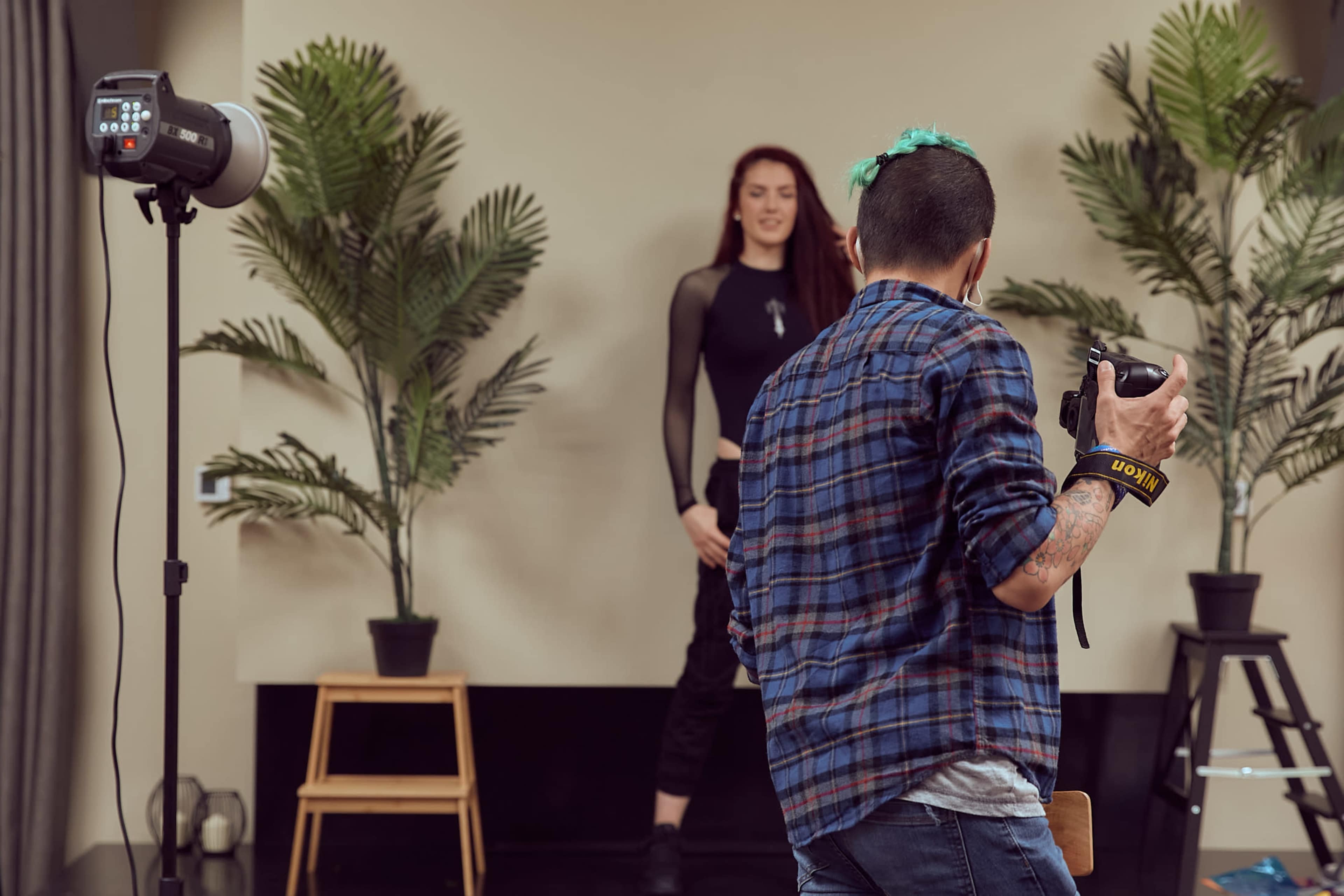 A person holding a camera is photographing a model posed in front of potted plants, with a studio lighting setup in the background.
