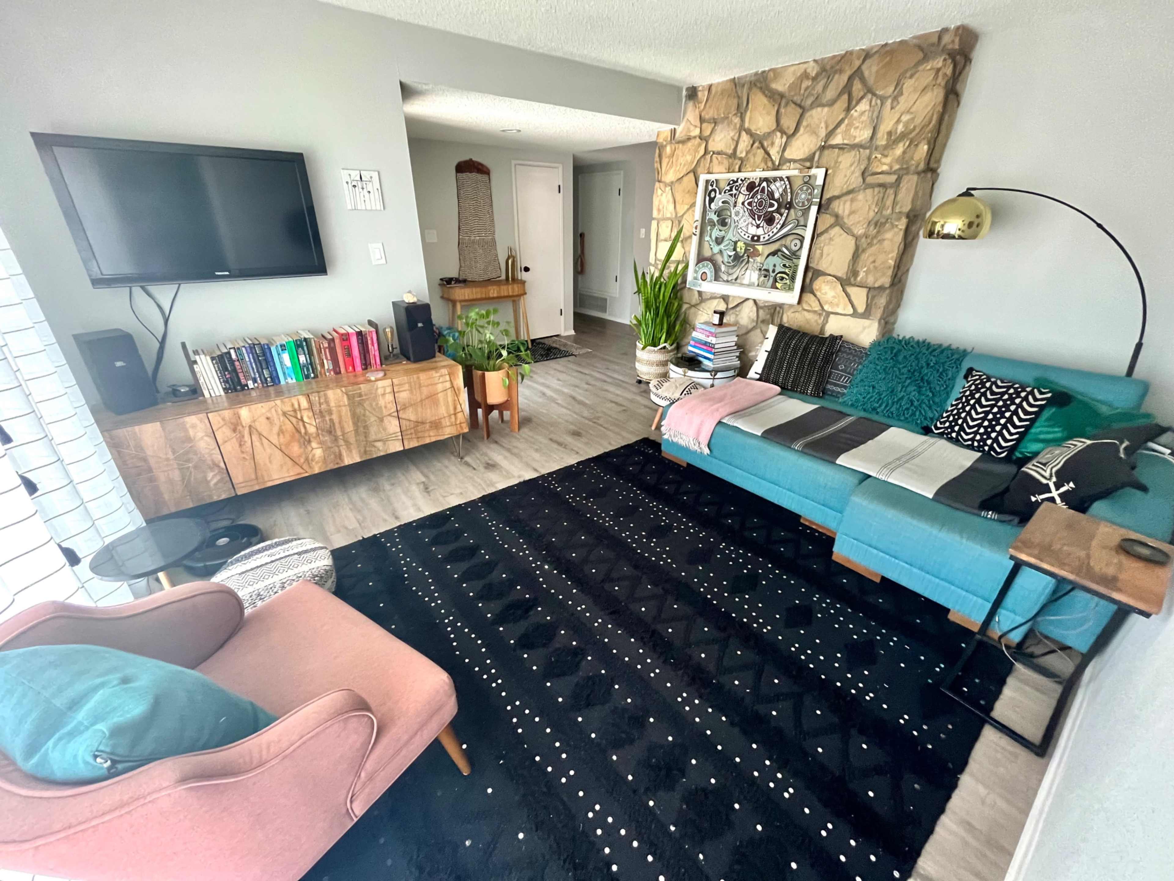 The living room features a blue sofa, a pink armchair, a stone wall, a wall-mounted TV, and a large black rug with white patterns.