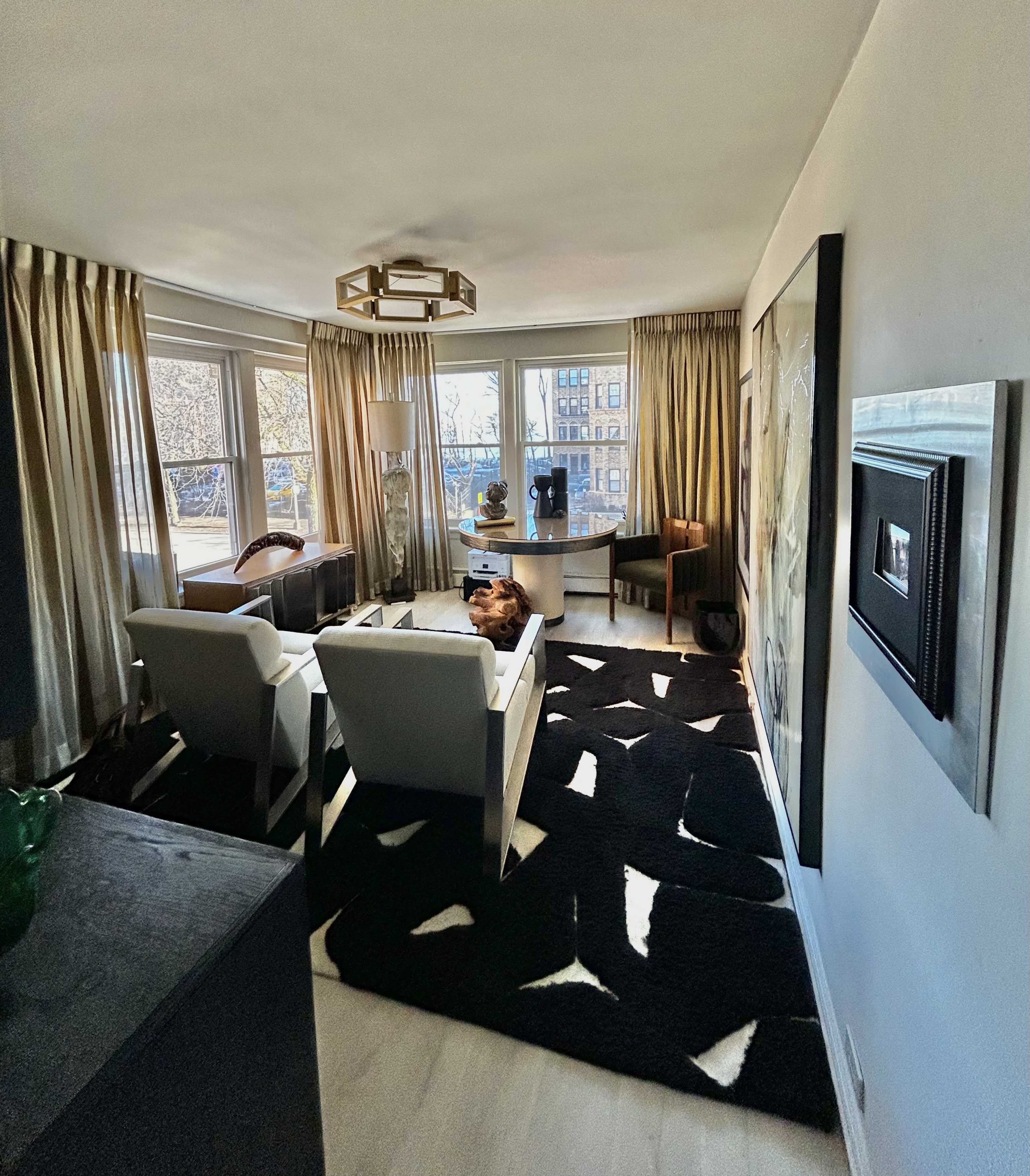 The image shows a modern living room with large windows, two white chairs, a black and white rug, and artwork on the walls.