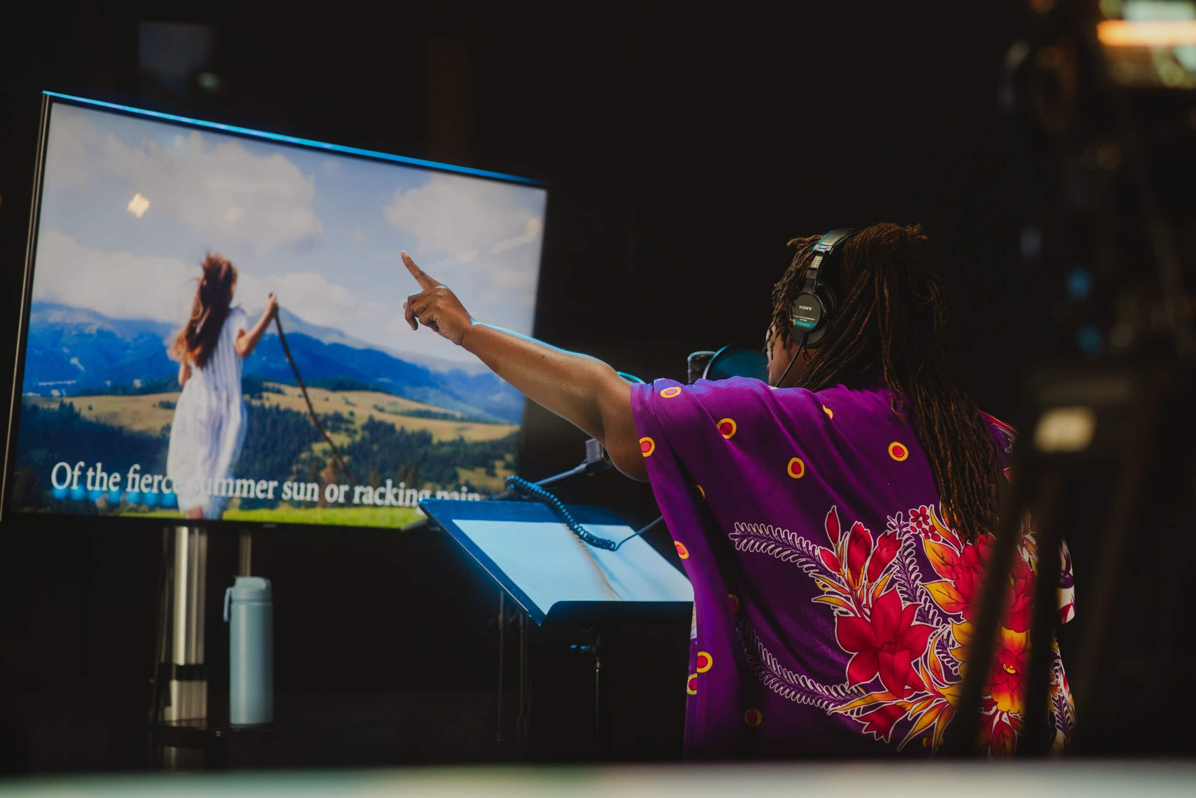 A person wearing a colorful shirt gestures towards a screen displaying a scenic landscape while participating in a recording session.