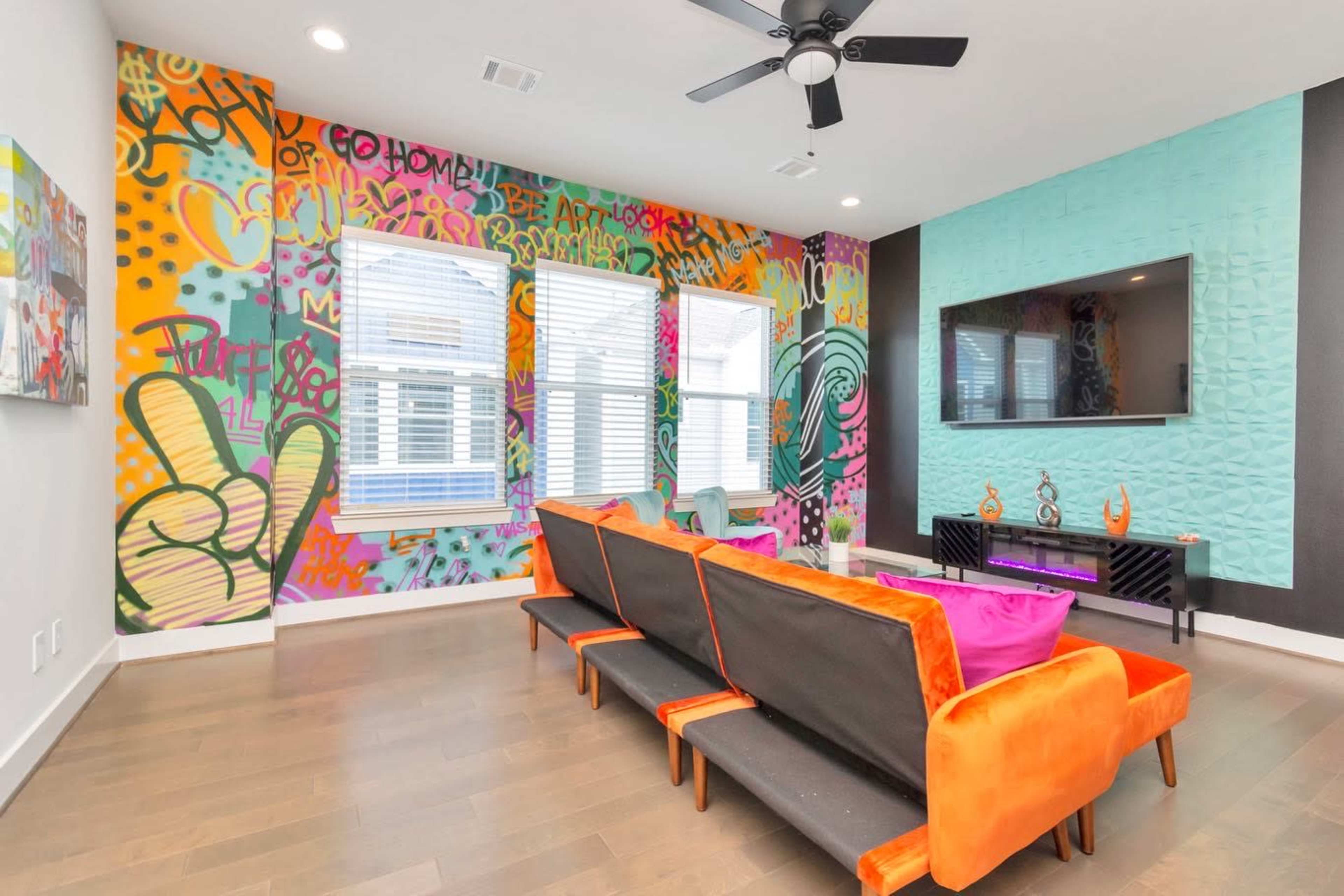 The room features a modern design with a colorful graffiti-style mural on one wall, large windows, and a black and orange sectional sofa facing a mounted television.