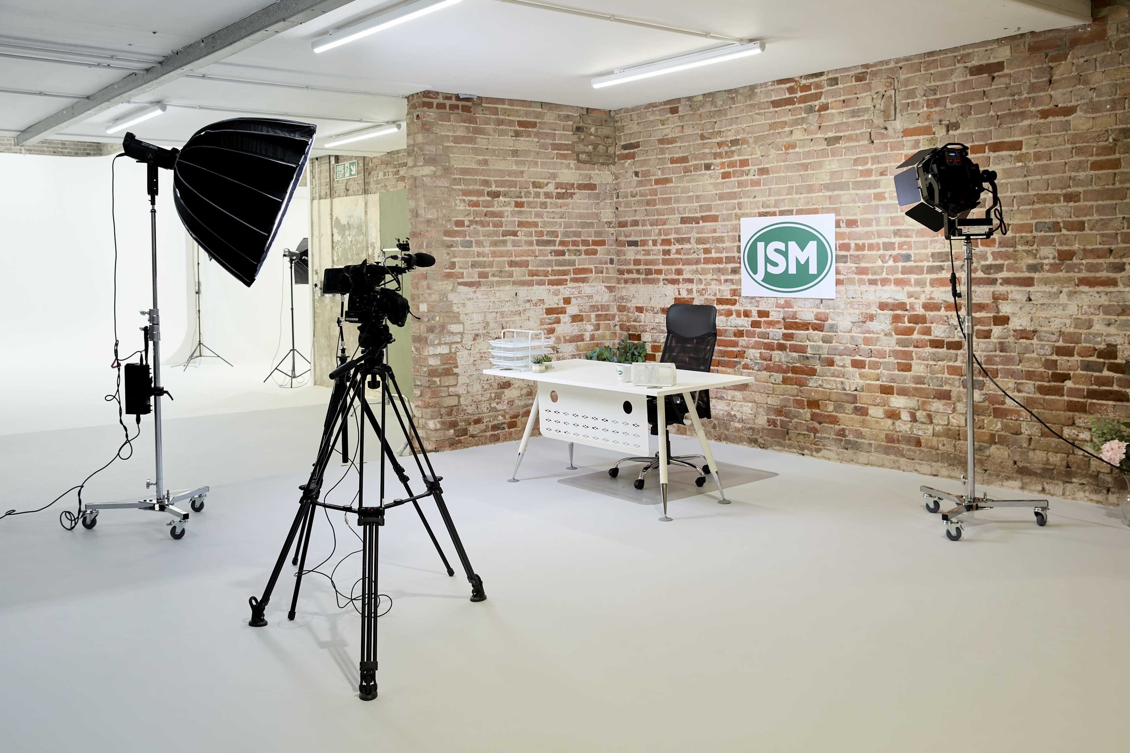 The image shows a studio setup featuring a desk with a chair, two cameras on tripods, and lighting equipment against a brick wall.