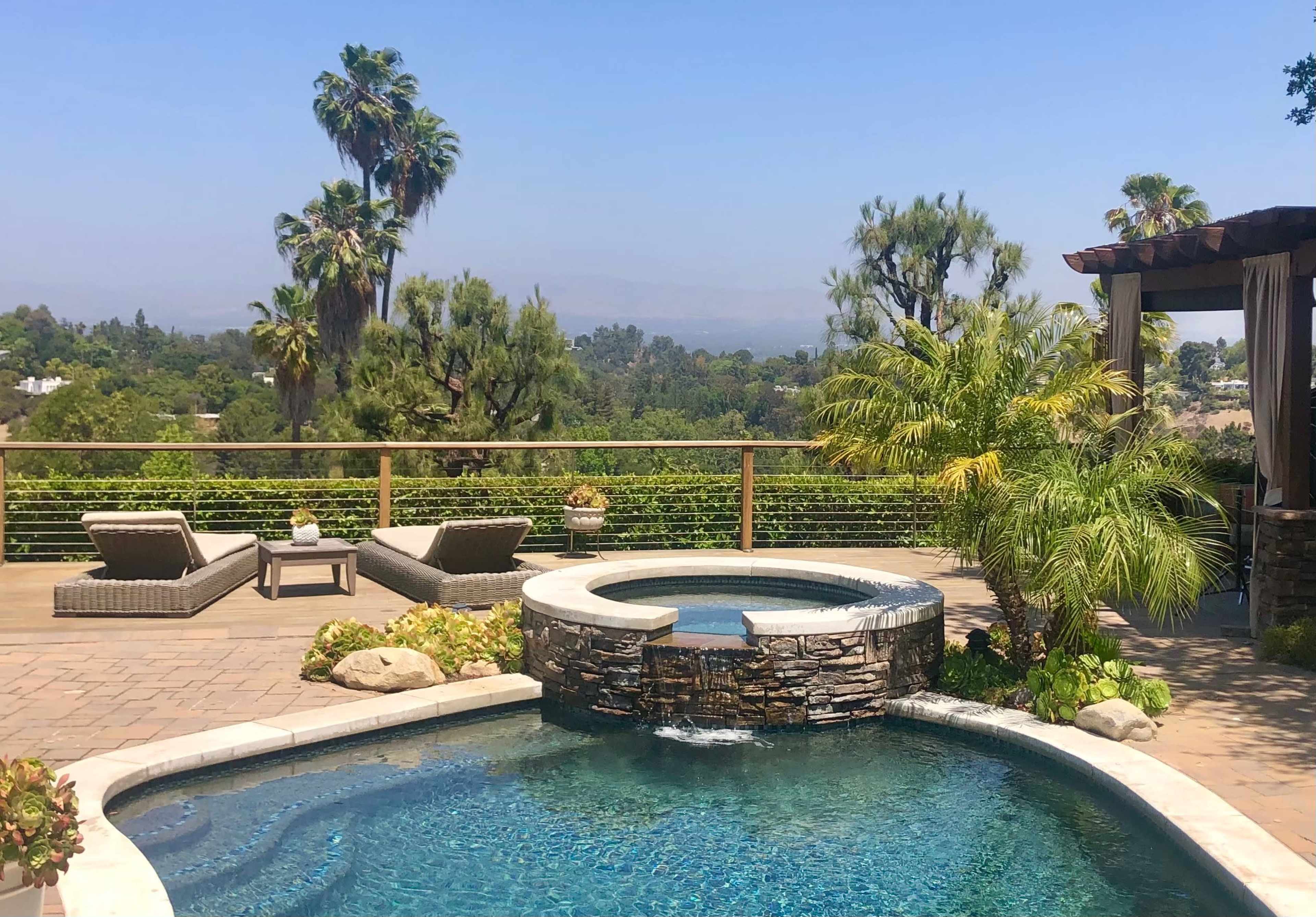 A landscaped backyard with a swimming pool featuring a stone waterfall and two lounge chairs overlooking a view of greenery and palm trees.