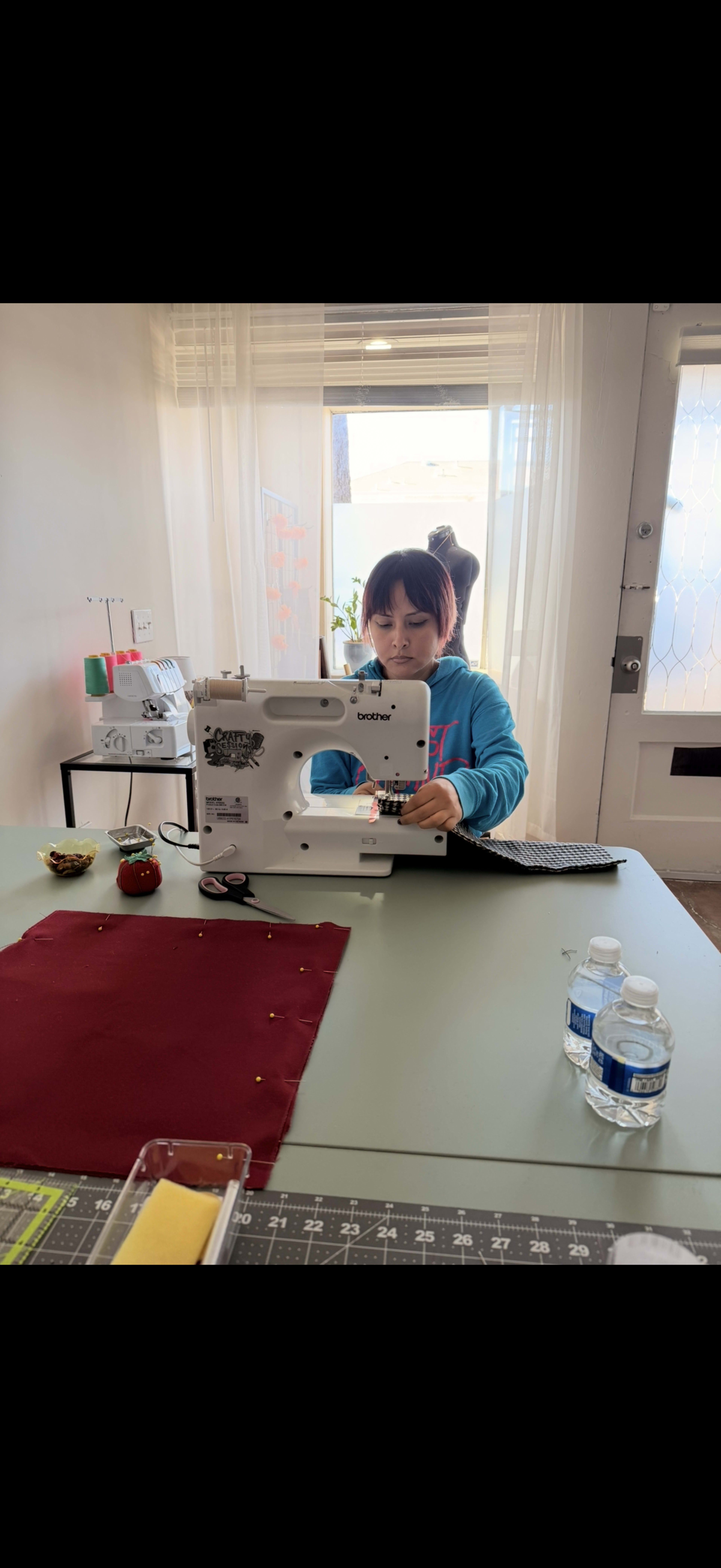 A child is using a sewing machine at a table with fabric, sewing supplies, and water bottles nearby.