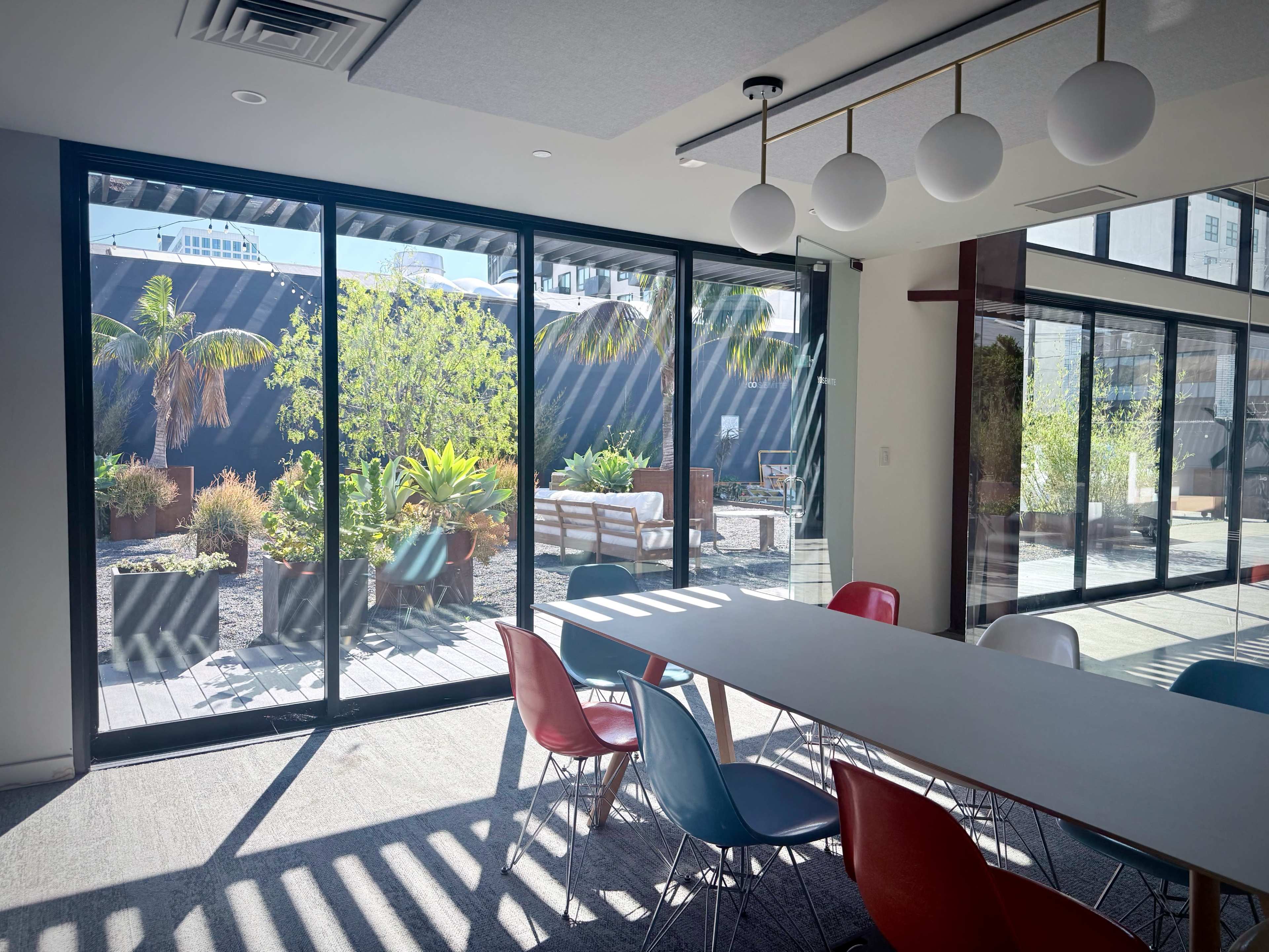 A bright meeting room features a large table surrounded by colorful chairs, with floor-to-ceiling windows showcasing a landscaped outdoor area.