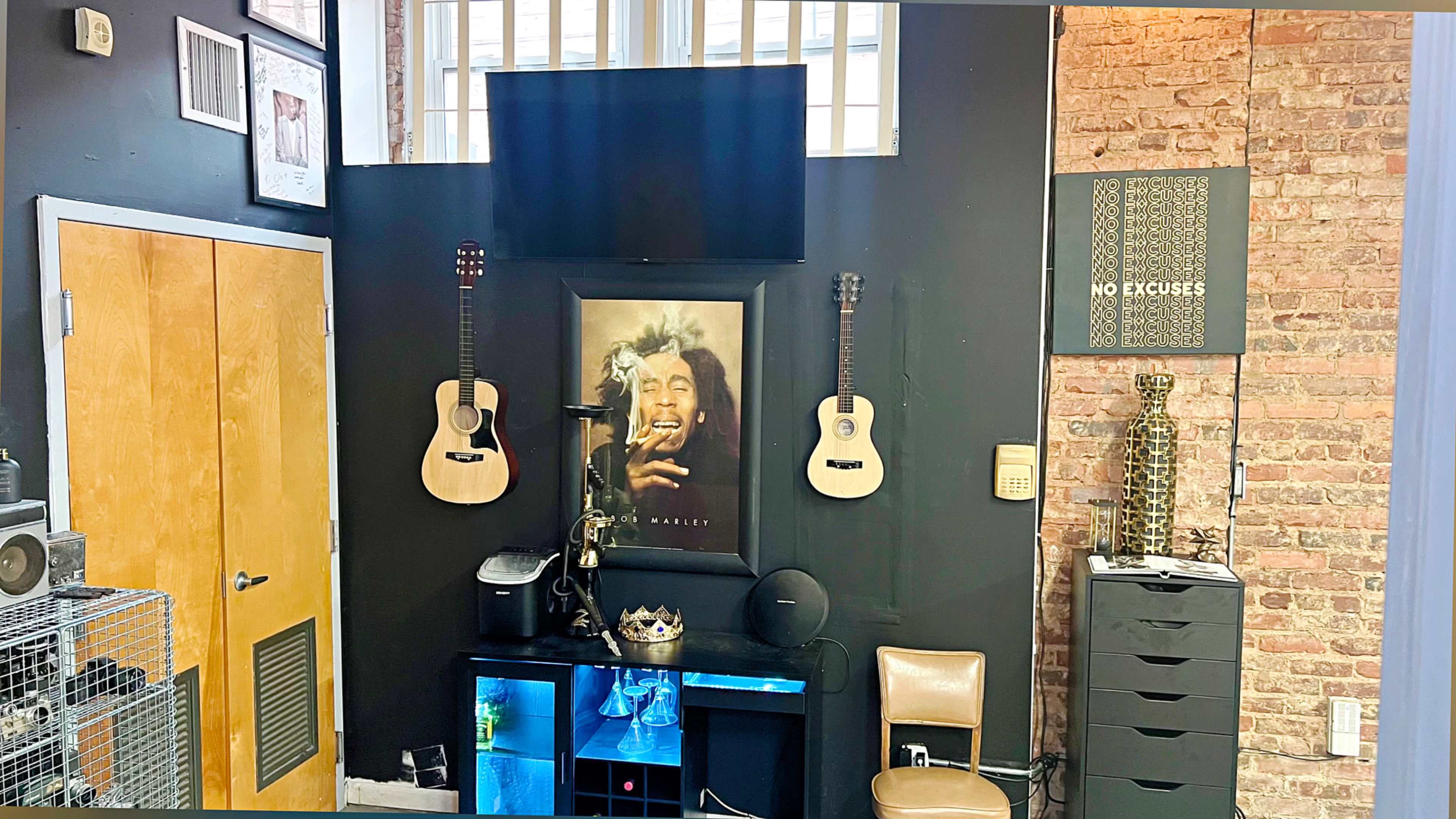 The image shows a room with a black wall featuring a framed portrait of a musician, two acoustic guitars mounted on either side, a small shelf with a crown on it, and a "No Excuses" sign on a nearby wall.