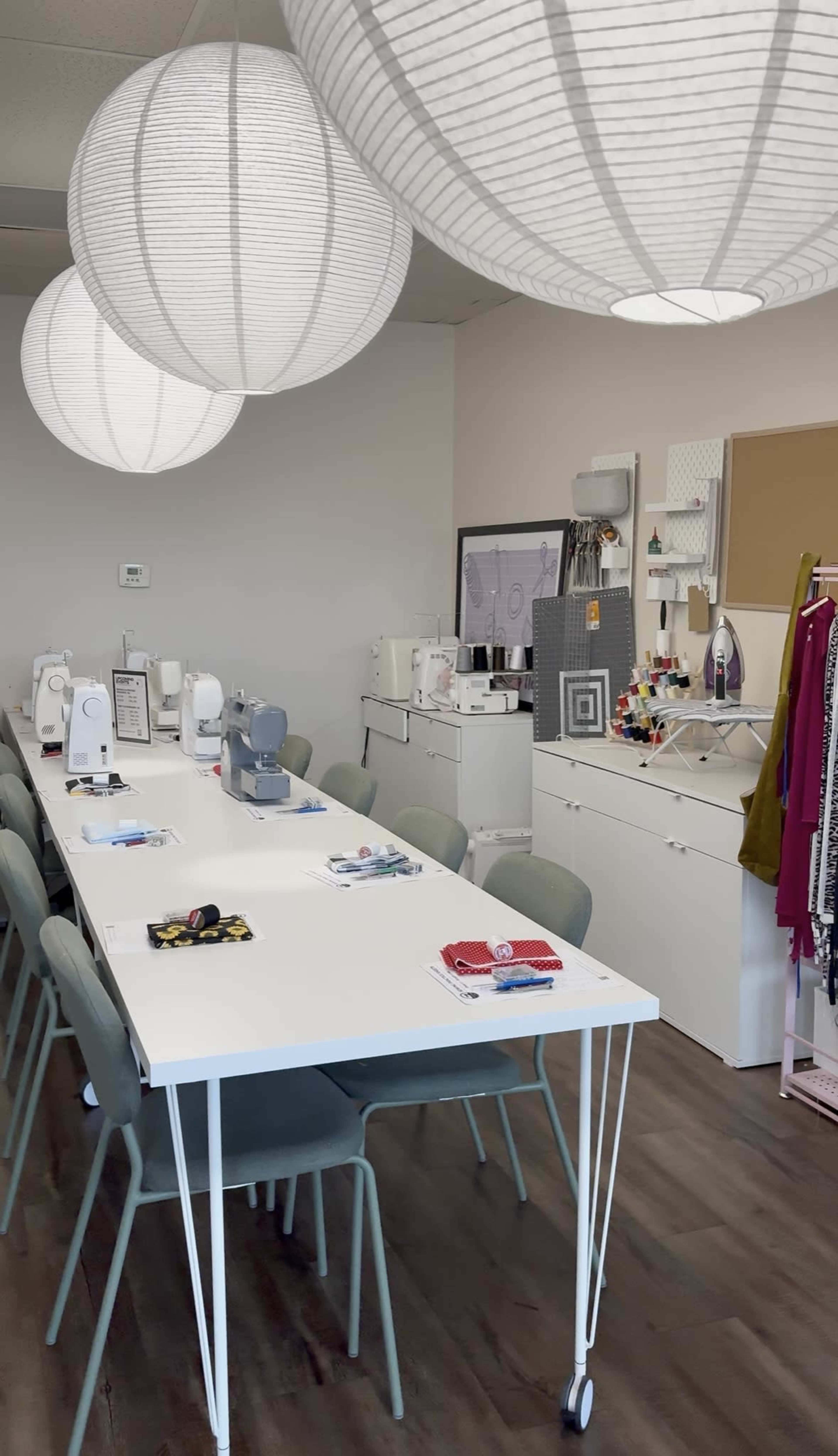 The image shows a spacious sewing studio with multiple sewing machines arranged on a long table, surrounded by light-colored chairs and adorned with large paper lanterns overhead.