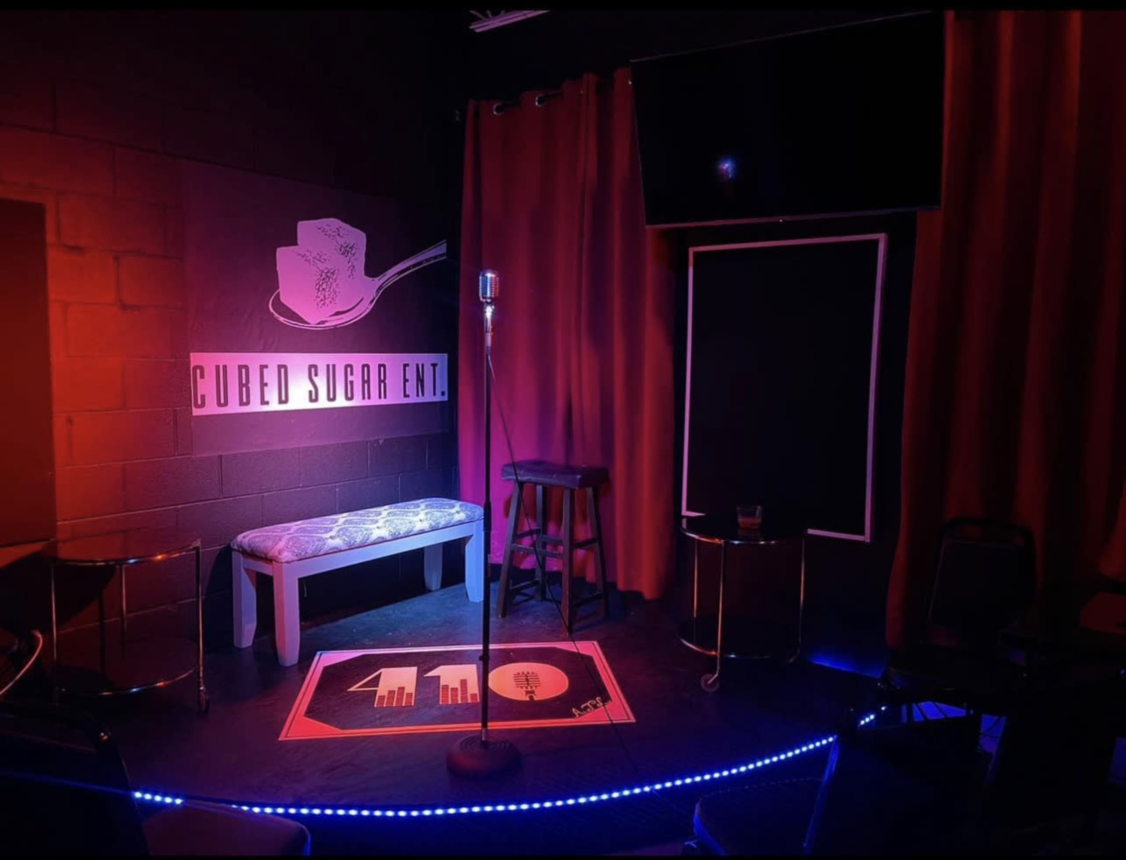The scene shows a small stage area featuring a microphone on a stand, a bench, and a bar stool, illuminated by colored lights with a sign reading "Cubed Sugar Ent." in the background.