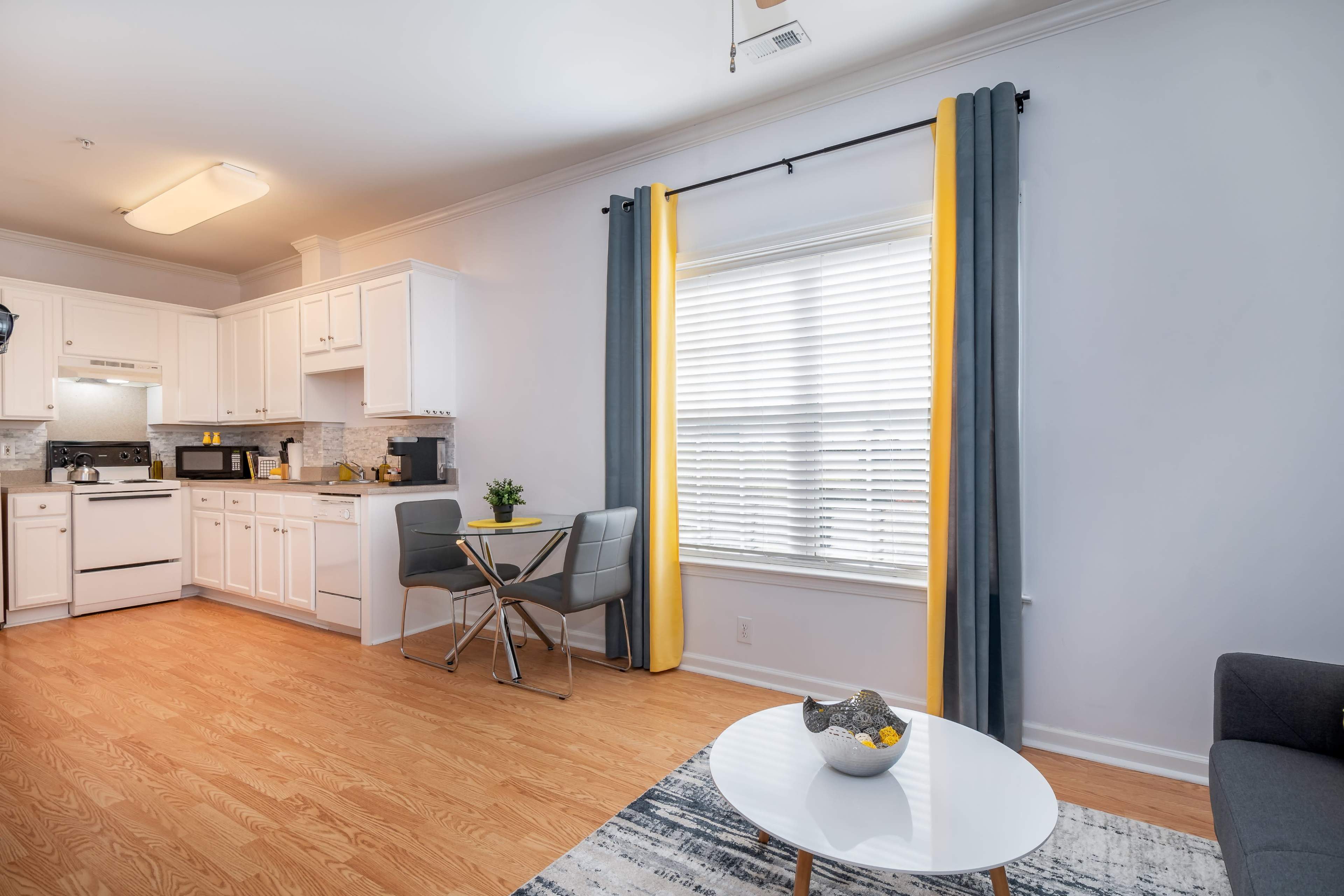A modern living space featuring a kitchen area with white cabinets, a dining table with two chairs, and a sofa, all illuminated by natural light through a window with yellow curtains.