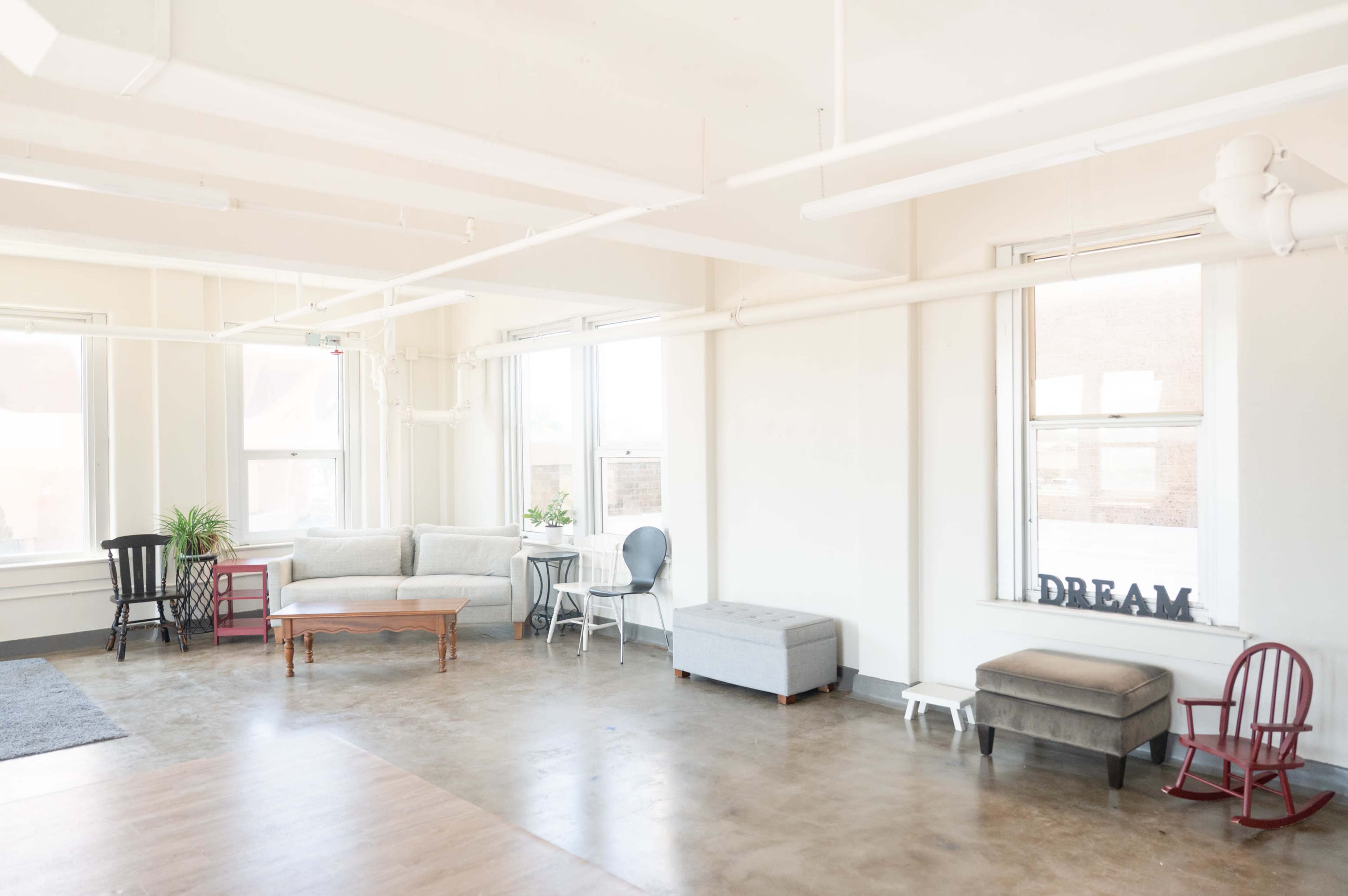 The image shows a spacious, modern room with hardwood and concrete floors, featuring various seating options, windows allowing natural light, and minimalistic decor.