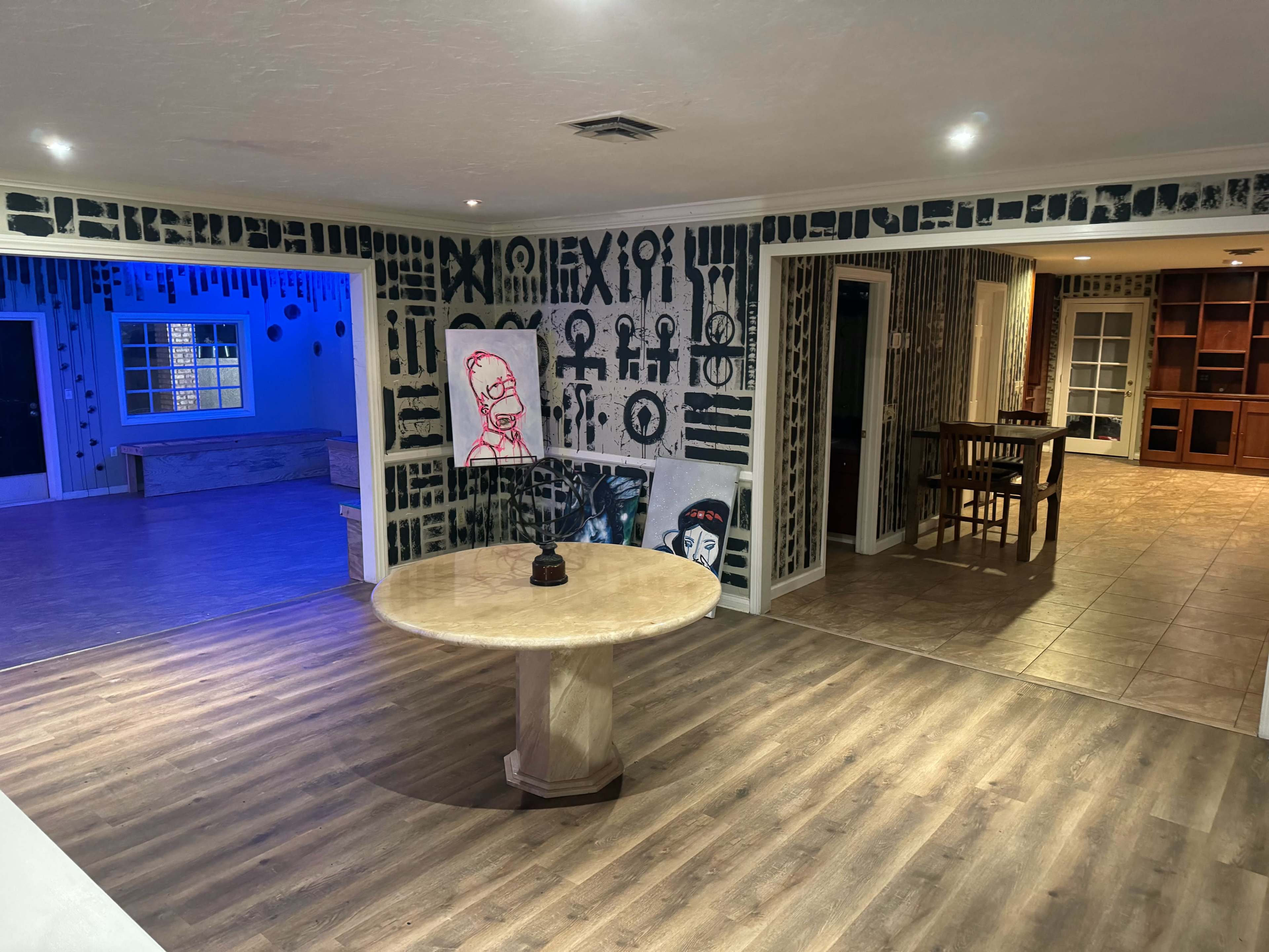 A spacious interior with distinct wall art featuring patterns, a round table in the foreground, and two adjacent rooms, one illuminated in blue.