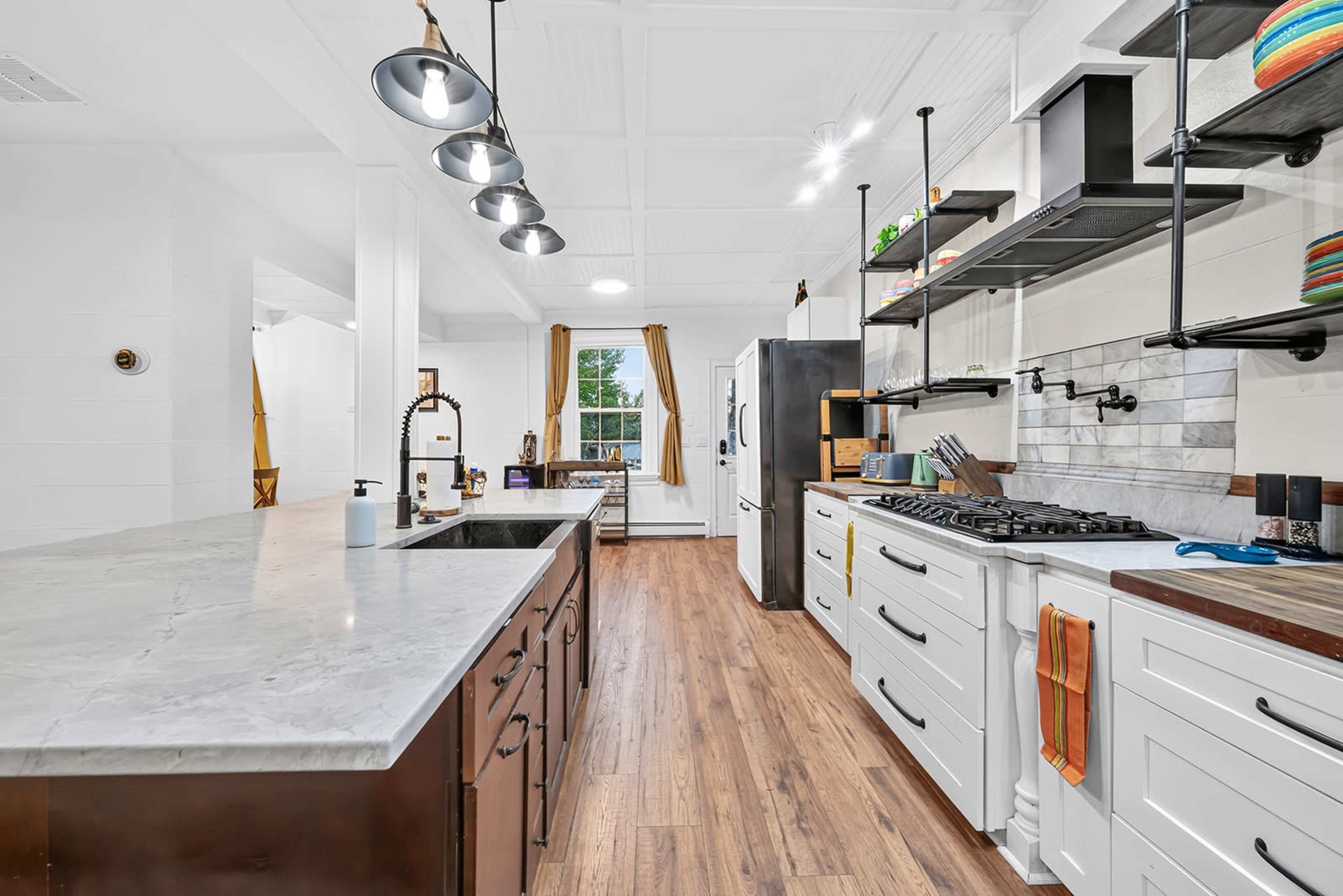 The image shows a modern kitchen with a large marble island, white cabinetry, and open shelving displaying colorful dishware.