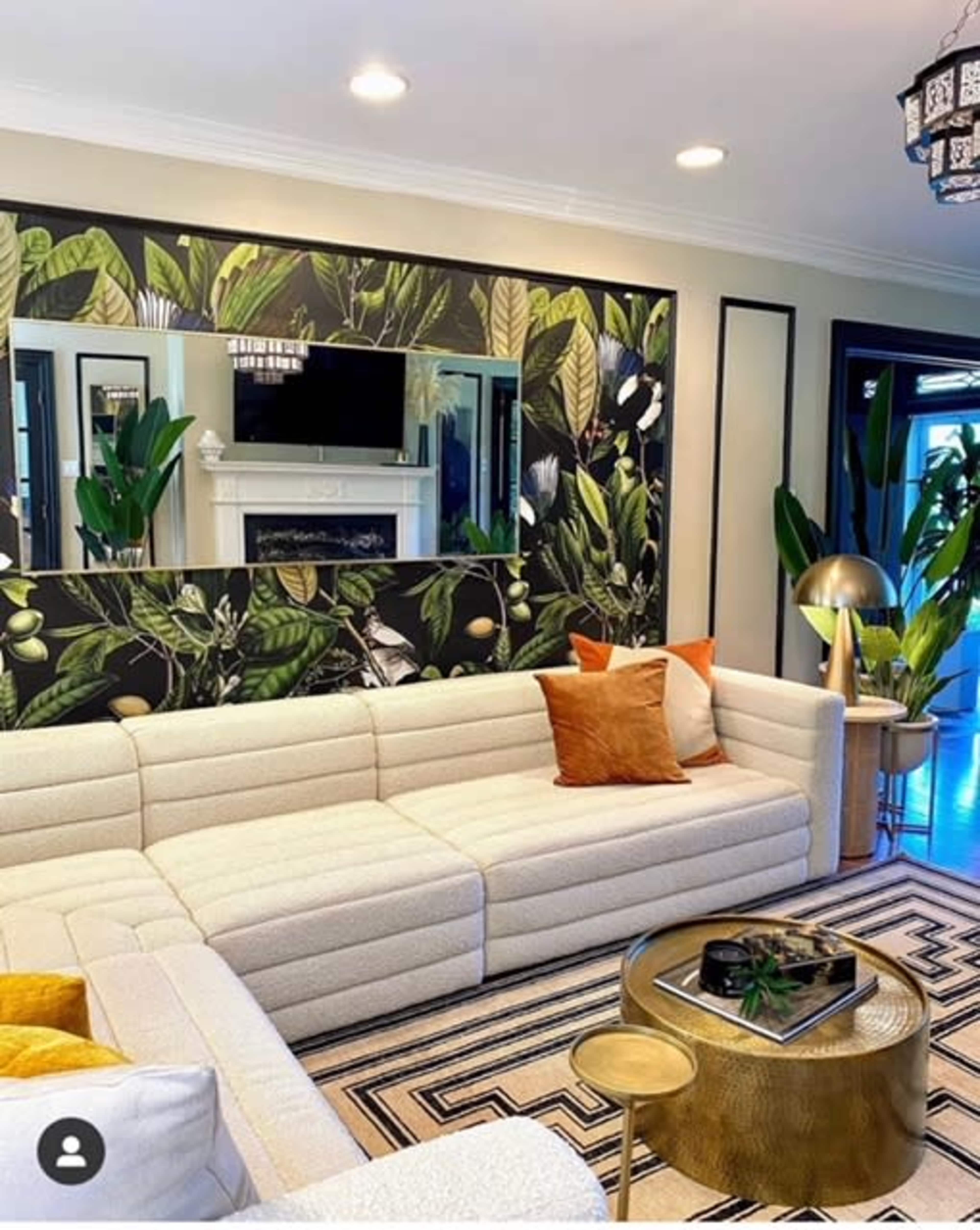 A stylish living room featuring a white sectional sofa, a patterned rug, a gold coffee table, and a wall adorned with a botanical mural.