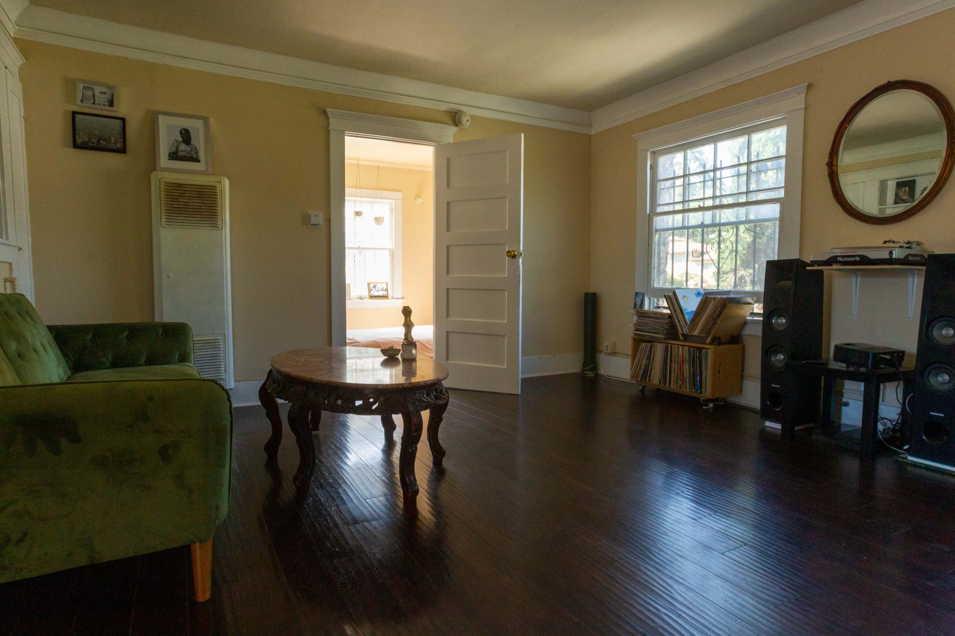 A living room with a green couch, a round wooden table, a wall of records, and an open door leading to a well-lit adjacent room.
