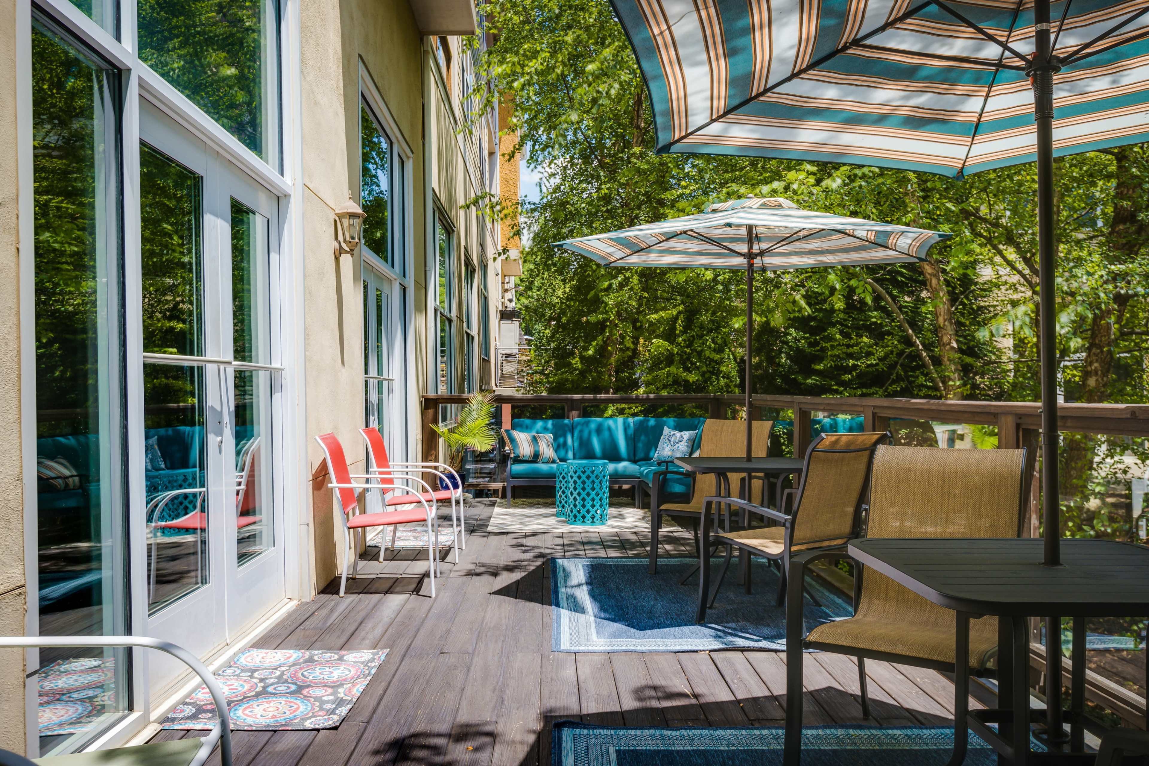 The image shows a sunlit outdoor patio with various seating arrangements, including chairs and a table, surrounded by greenery and umbrellas.