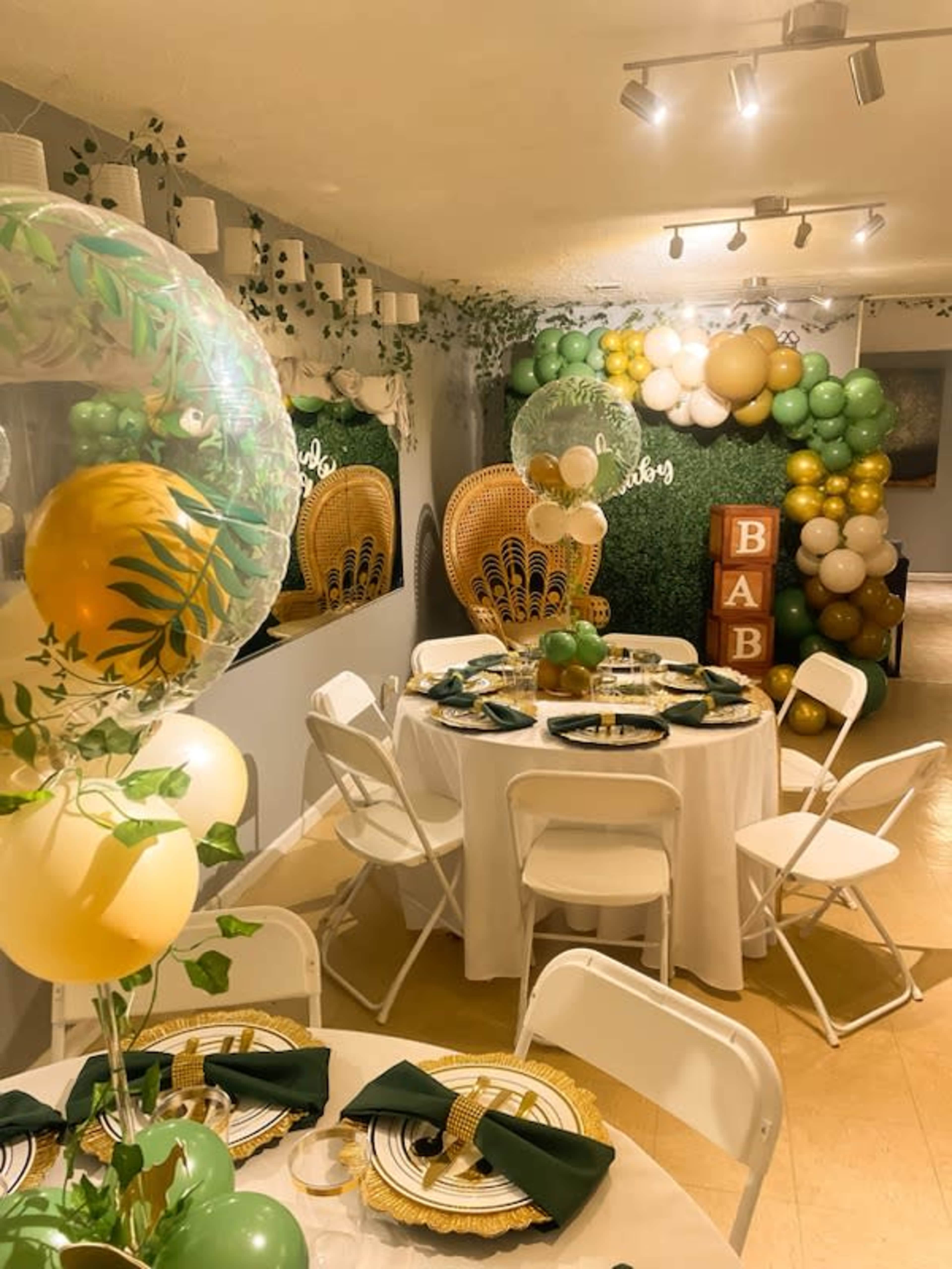 The image displays a decorated party space with a round table set for a celebration, featuring elegant tableware, gold and green balloons, and a backdrop with foliage-themed decor.