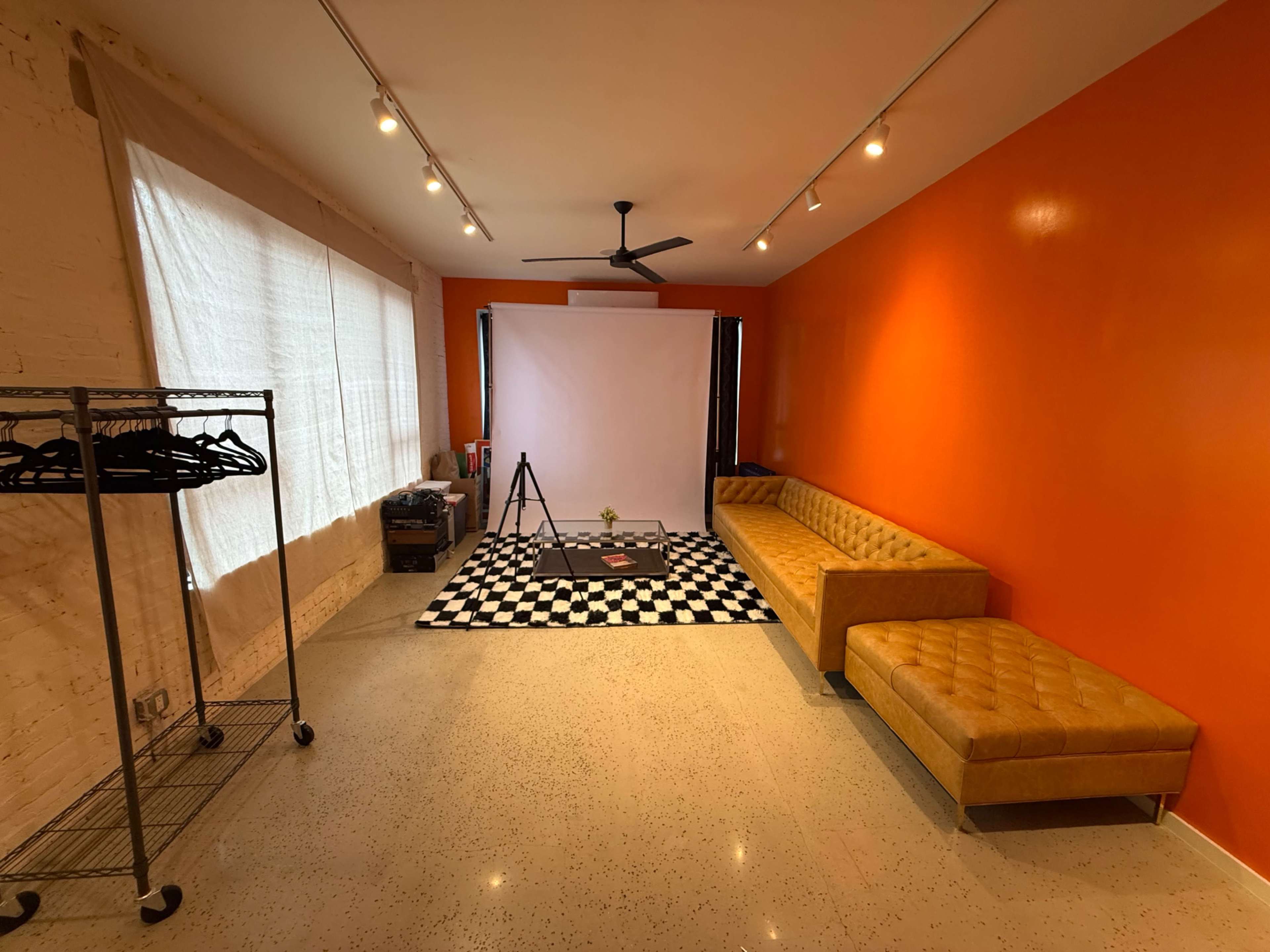 The image shows a spacious room with an orange accent wall, a black and white checkered rug in the center, a tripod set up in front of a blank backdrop, and a yellow sofa along one side.
