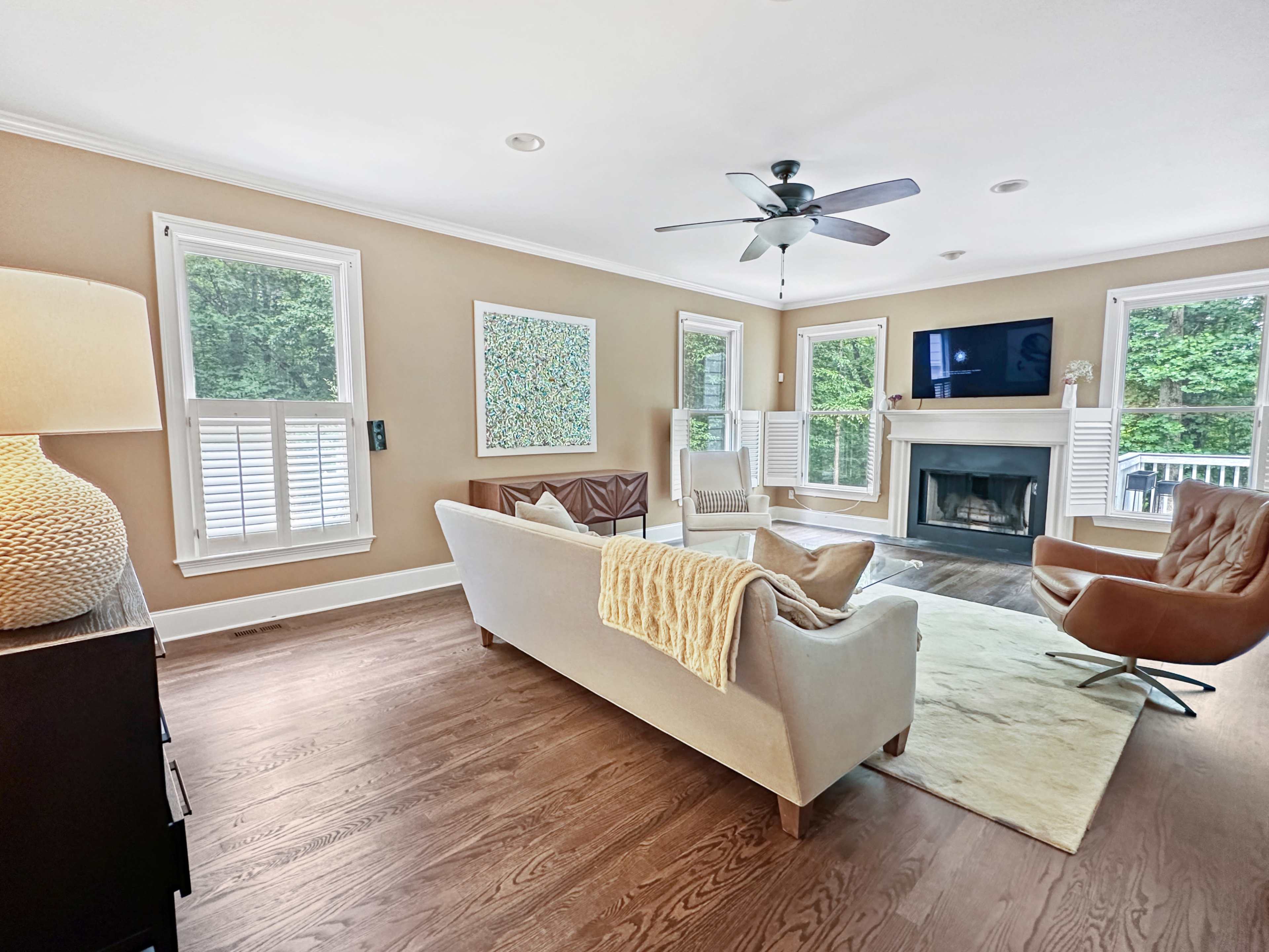 The bright living room features a beige sofa, a brown armchair, a fireplace, and large windows overlooking a green landscape.