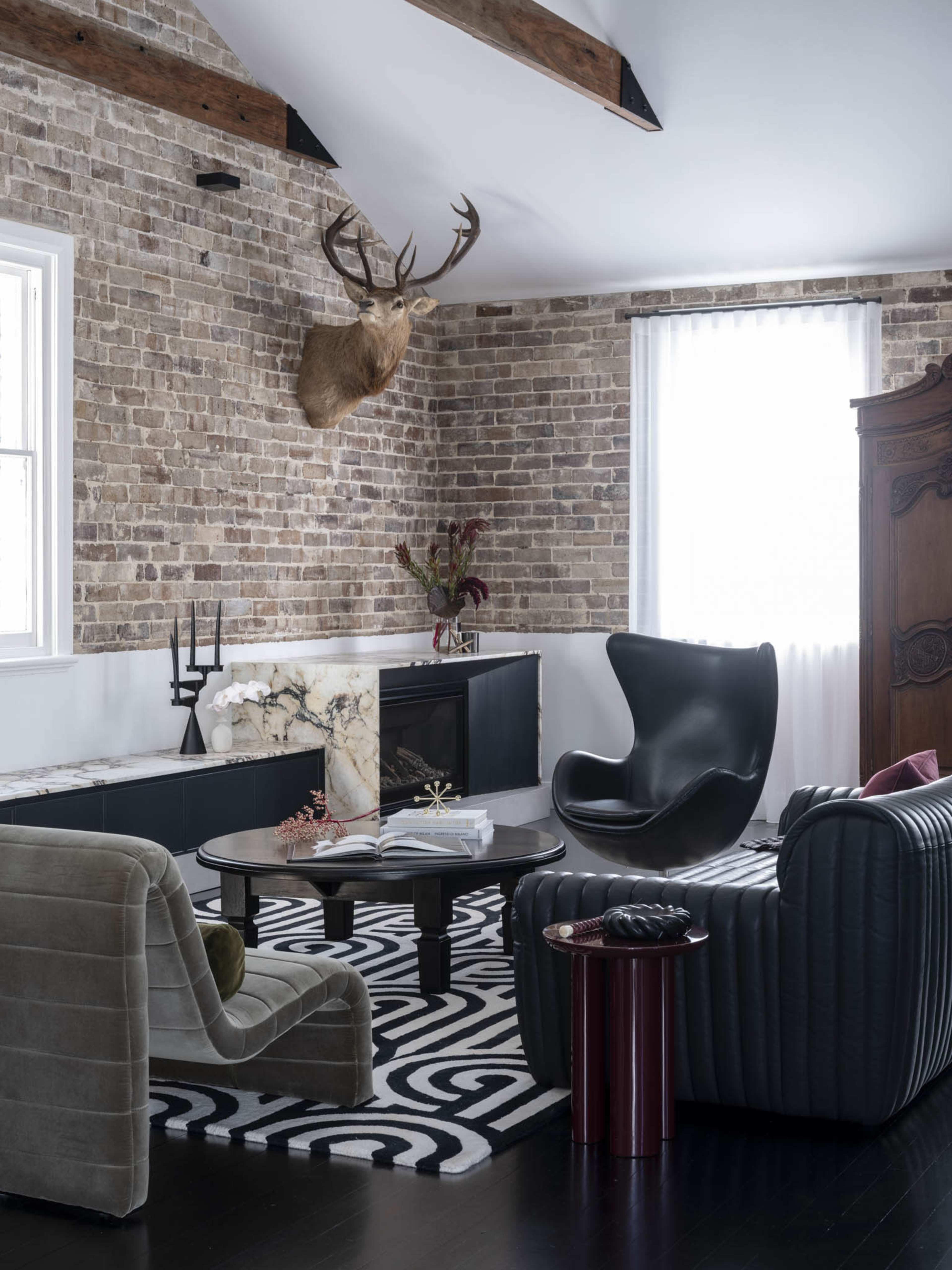 The image features a stylish living room with exposed brick walls, a mounted deer head, and a mix of modern furniture including curved chairs and a round coffee table on a patterned rug.