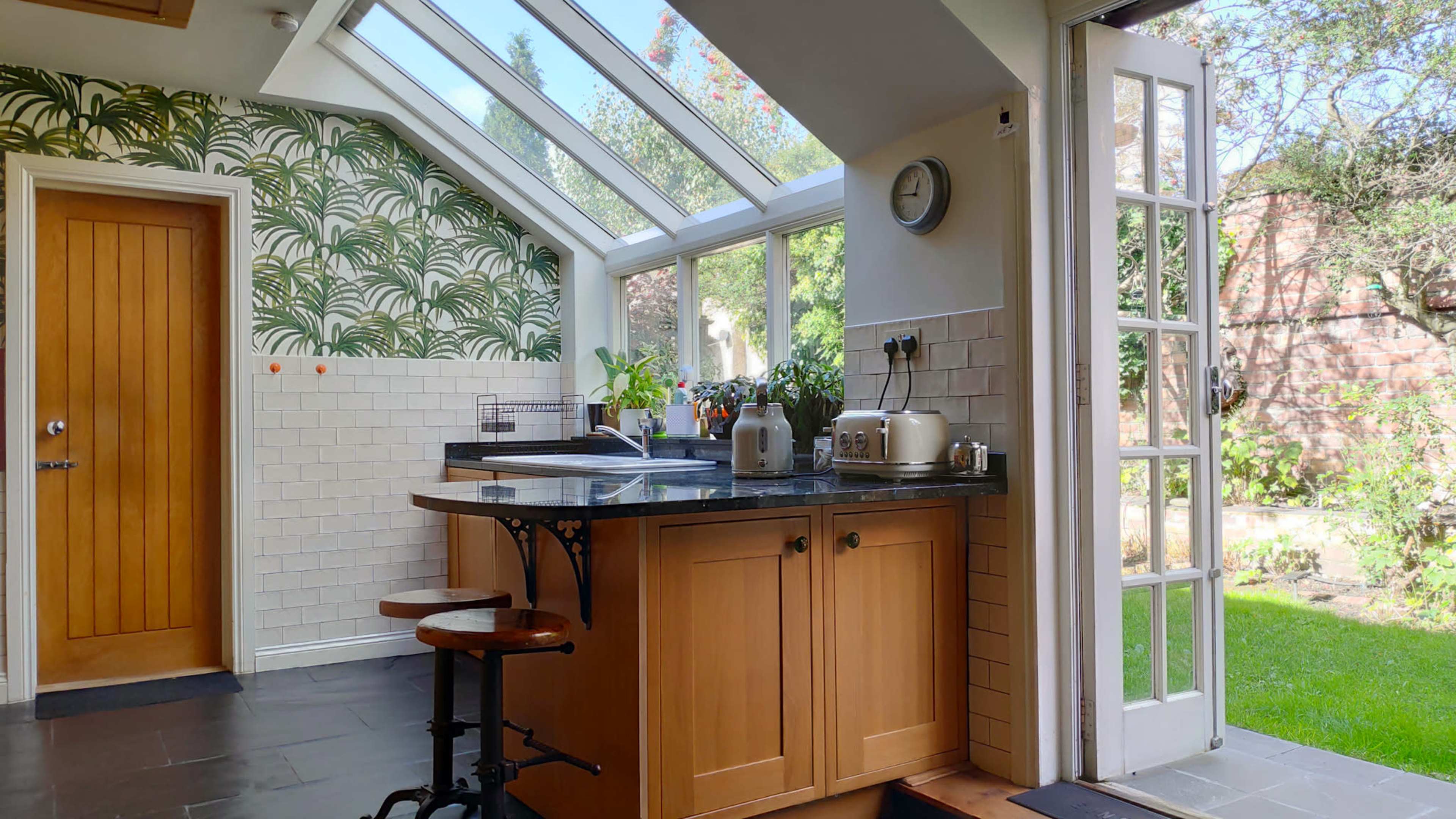 A kitchen with large windows, a wooden countertop, and a glimpse of a garden through an open door.