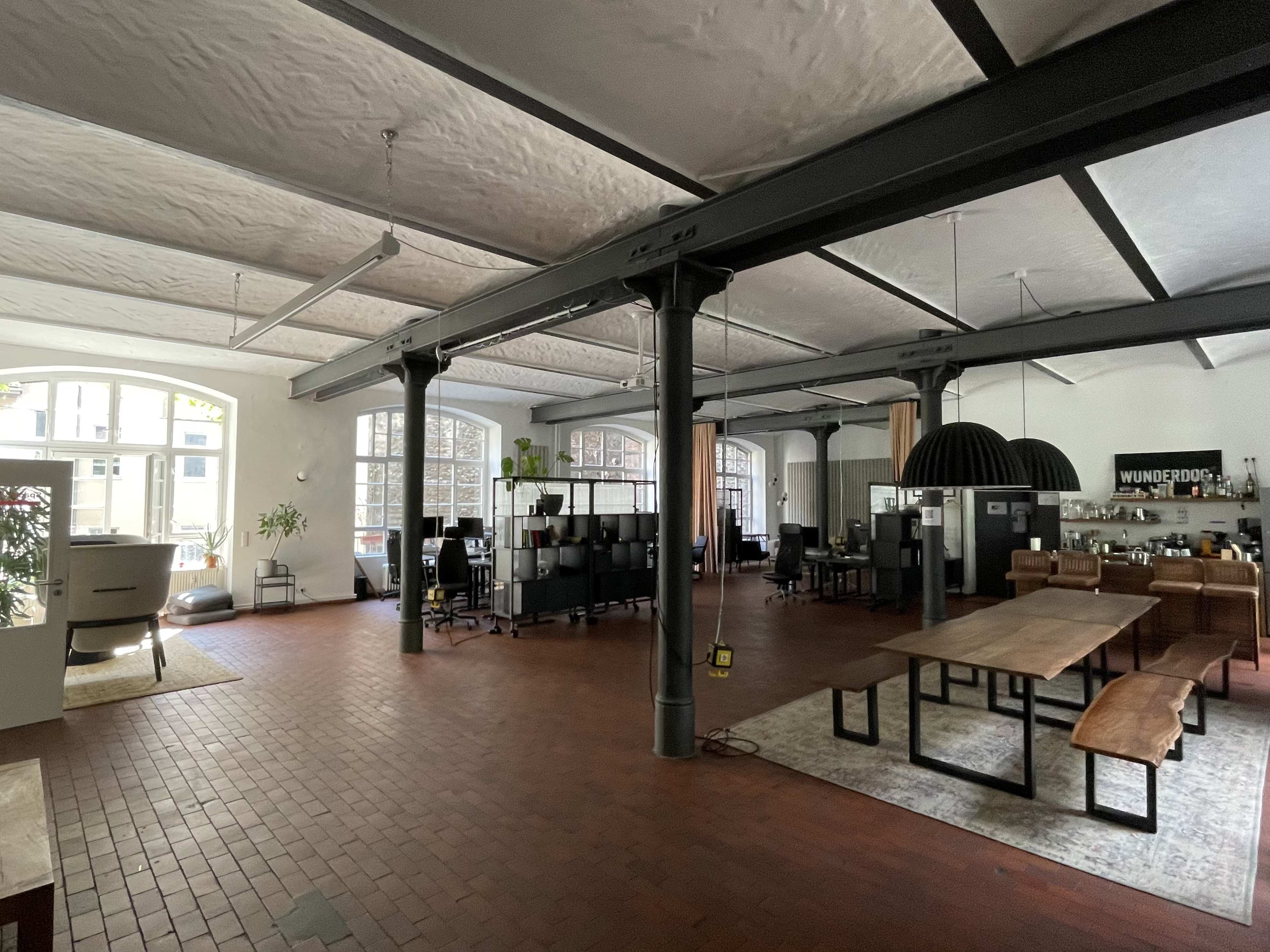 The image shows a spacious open office area with large windows, wooden furniture, and industrial-style support beams.