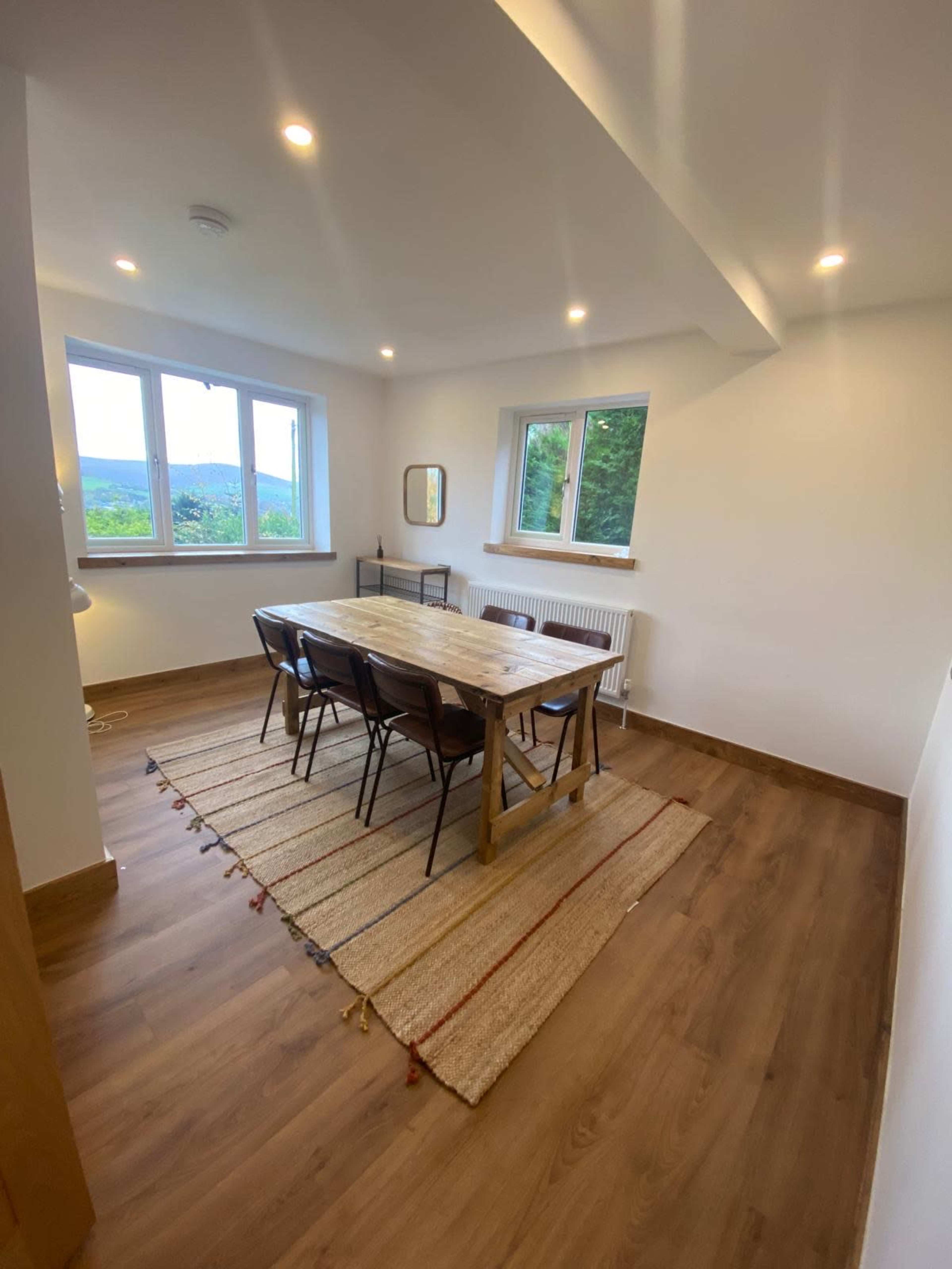 The image shows a dining area with a wooden table surrounded by six chairs, set on a textured rug, next to large windows that provide a view of greenery outside.