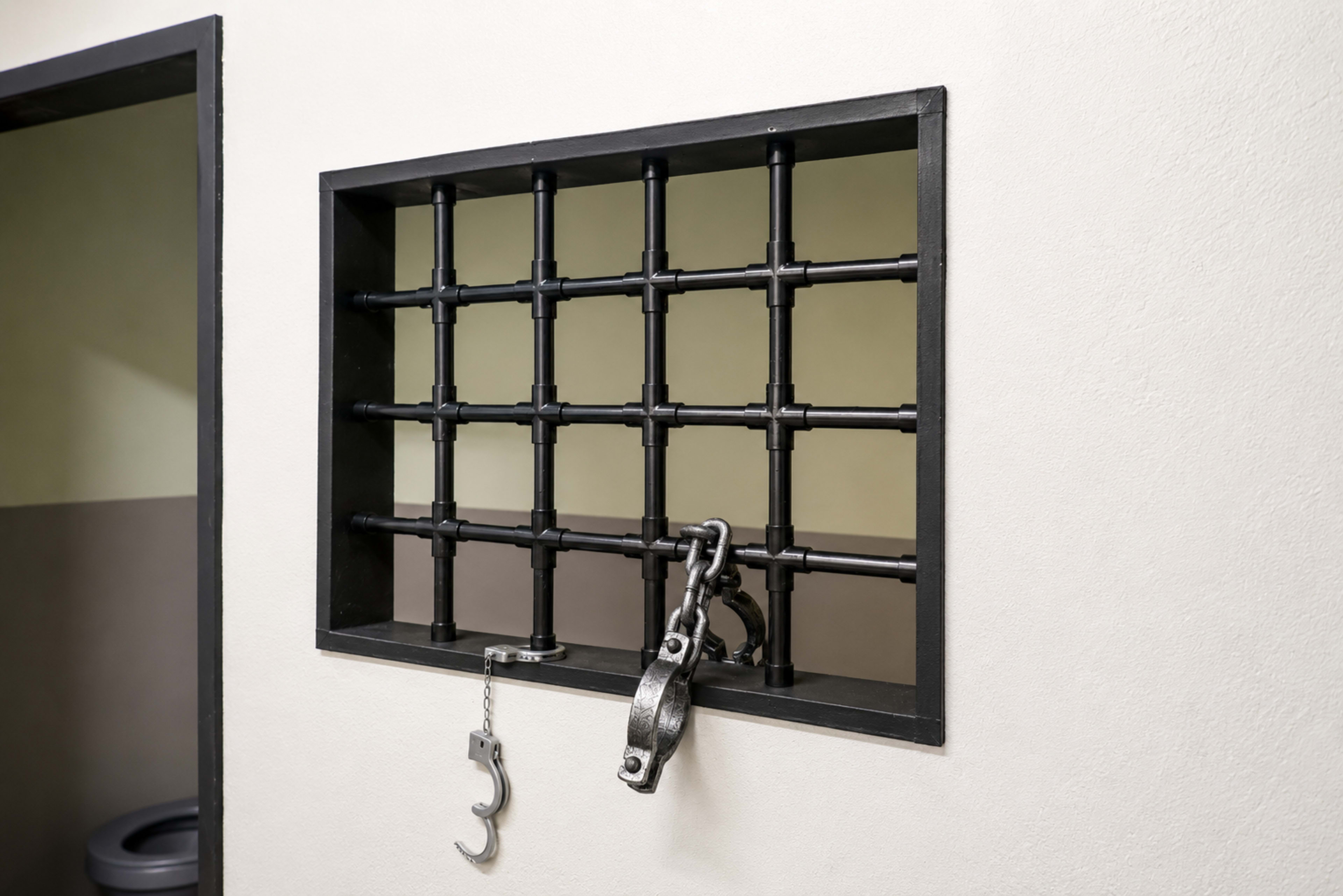 The image shows a barred window with a chain and hook attached, set into a wall in what appears to be a jail or prison.