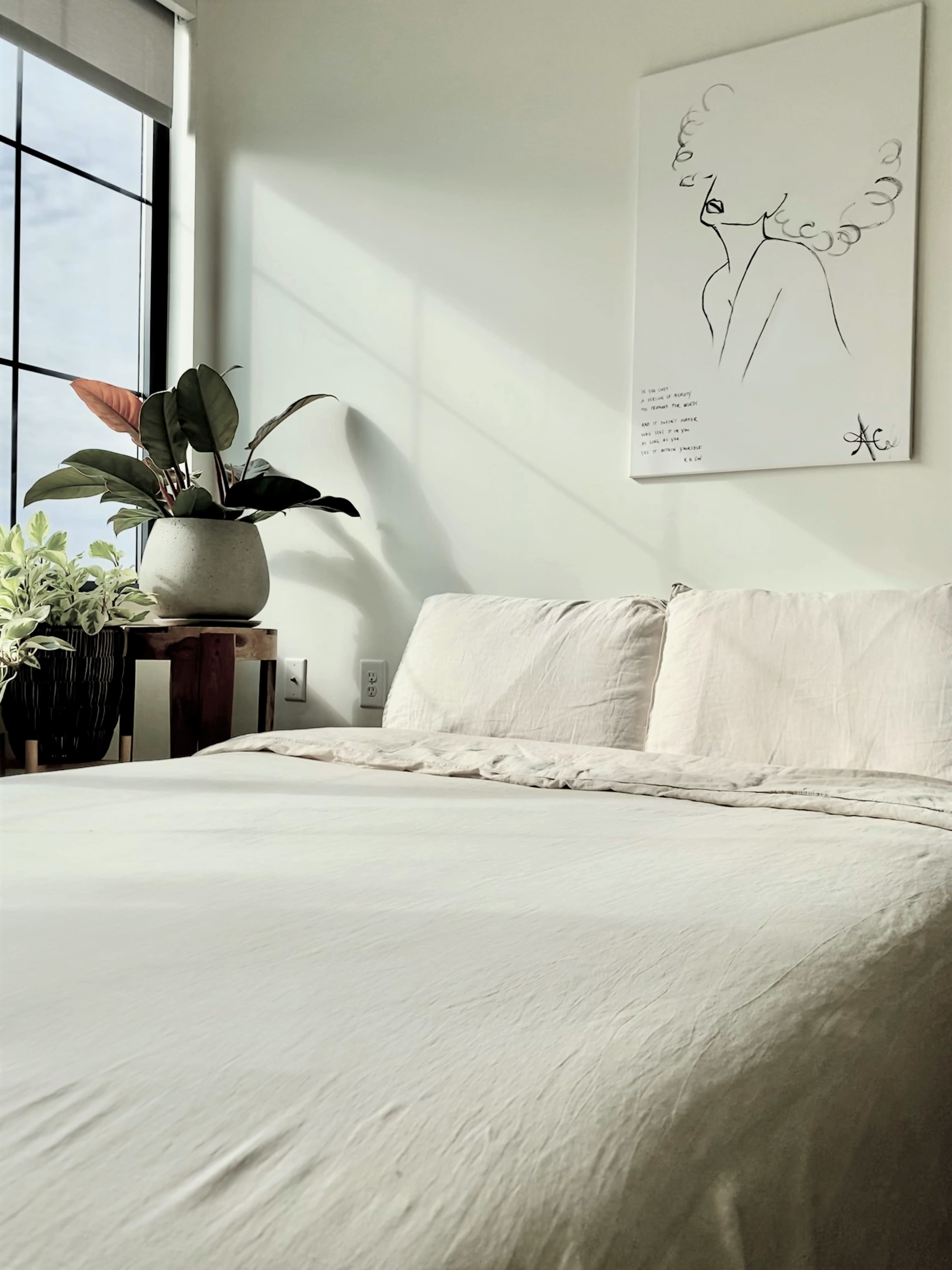 A neatly made bed with light-colored linens is positioned against a wall, accompanied by a potted plant and a framed artwork nearby.