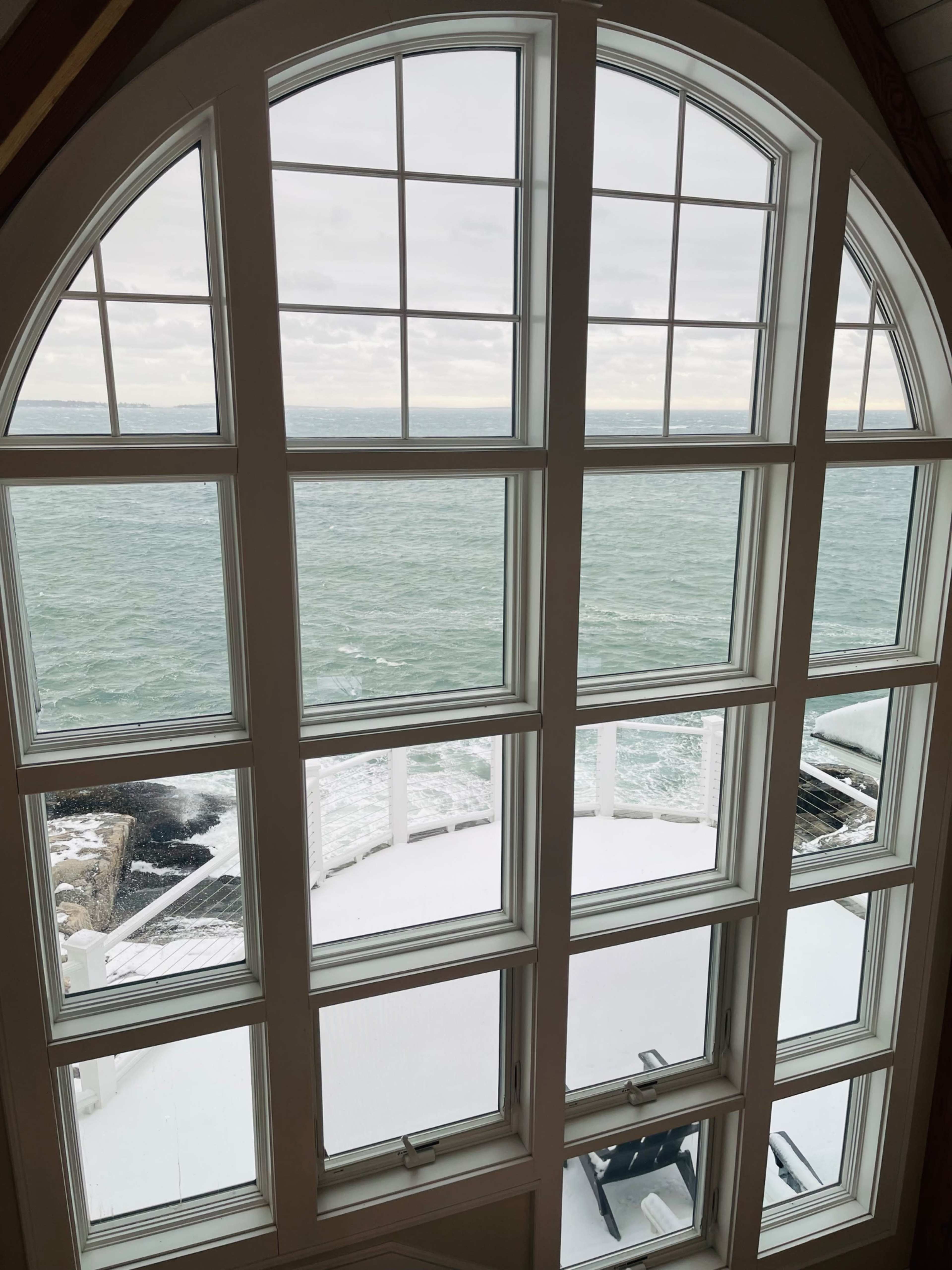The image shows a large arched window overlooking a snowy terrace and the ocean waves below.