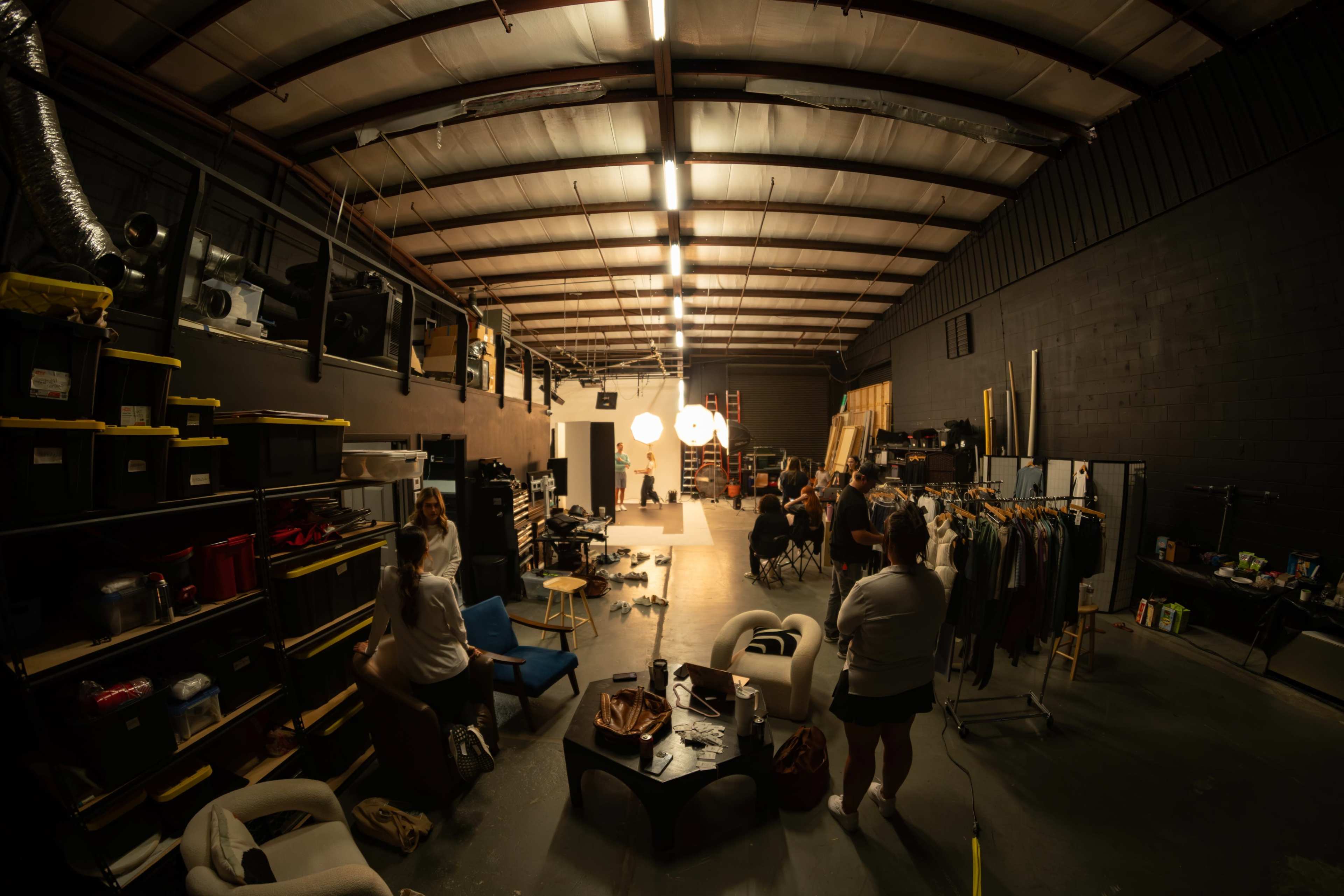The image shows a spacious studio environment with various storage units, clothing racks, and lighting fixtures arranged around a central area where a subject is being filmed.