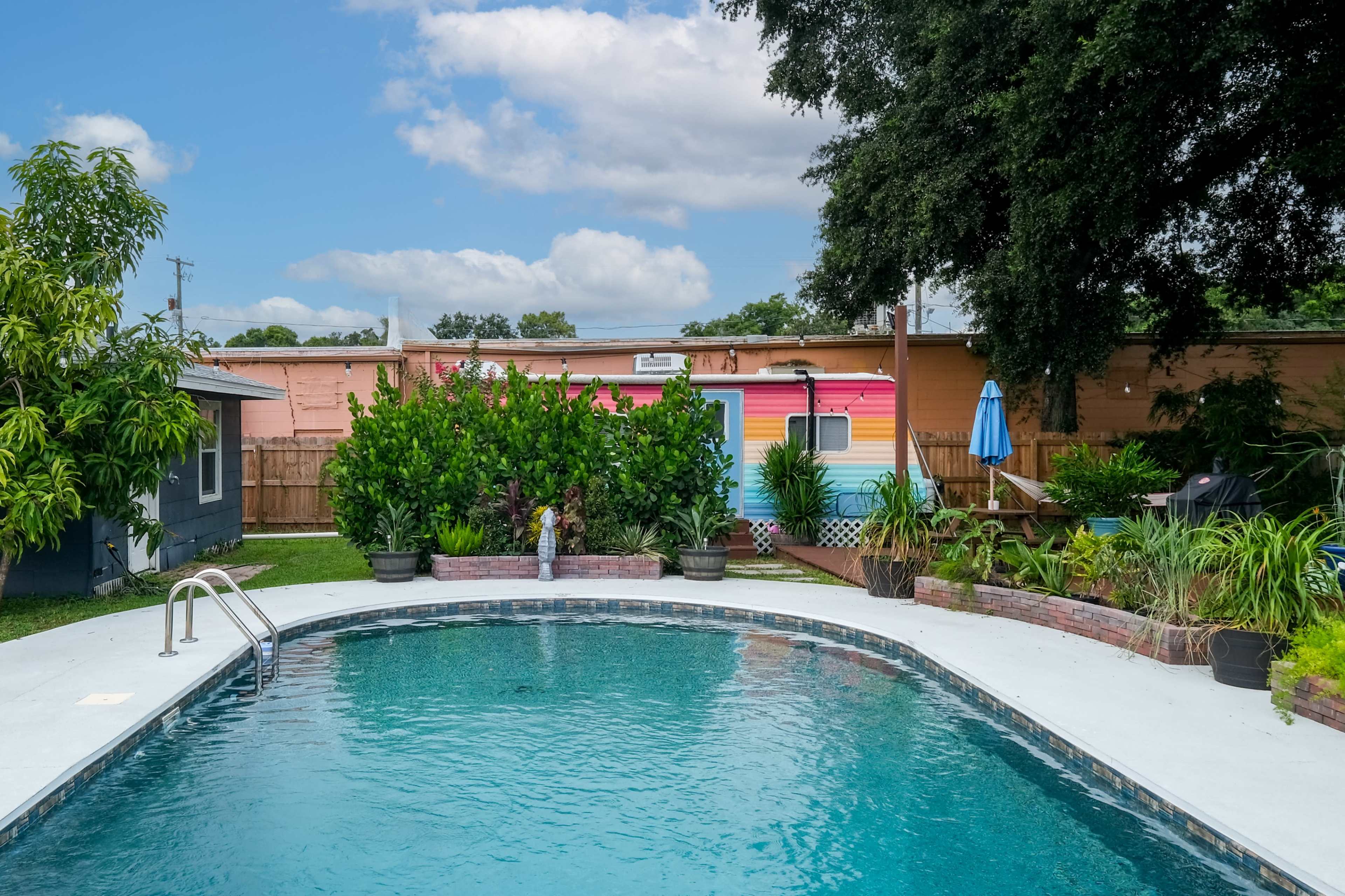 The image shows a swimming pool surrounded by landscaped greenery and colorful walls of a nearby building under a blue sky.