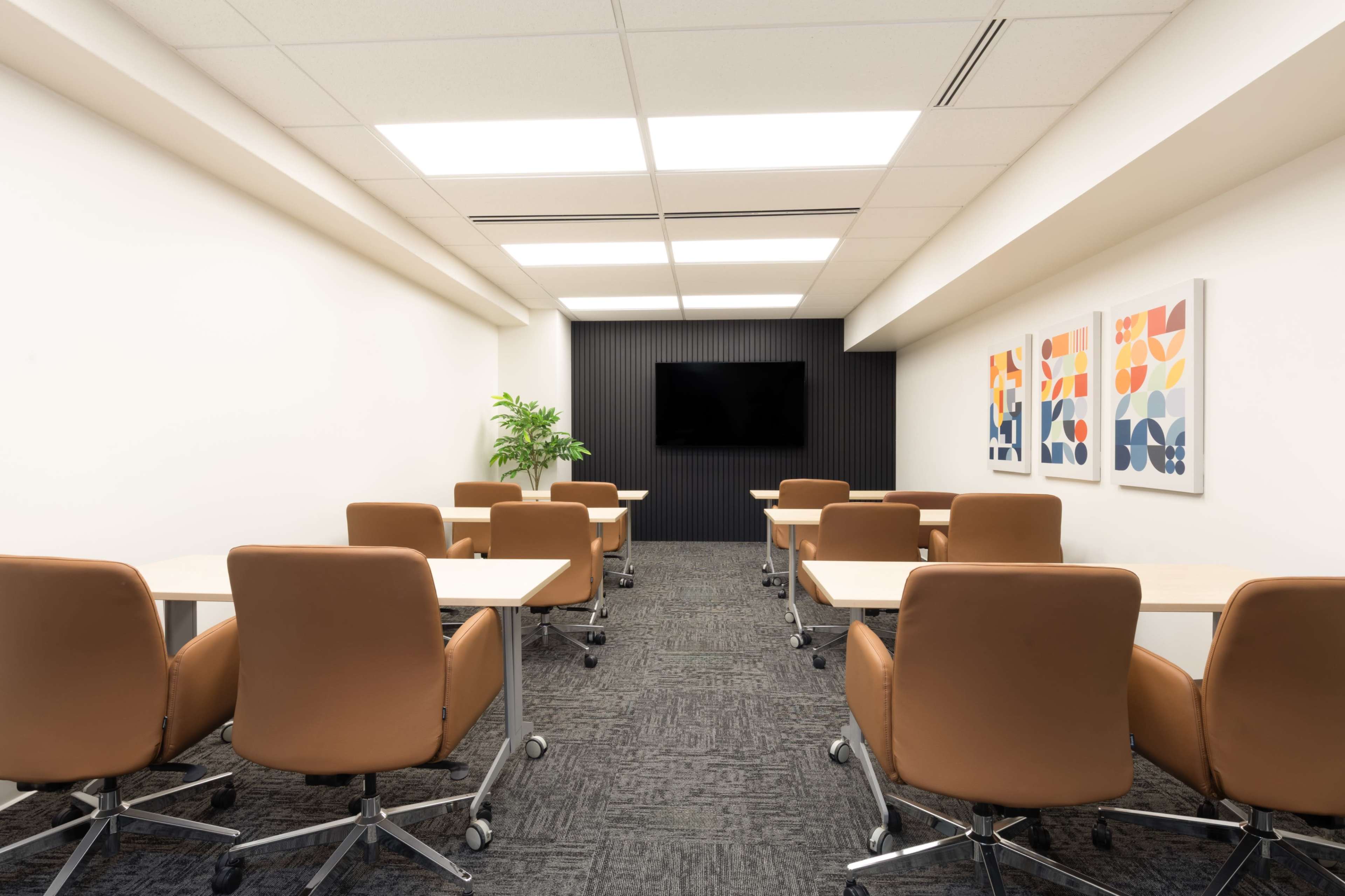 A modern conference room features multiple tan chairs with wheels and wooden tables arranged in rows, along with artwork on the wall and a television on a black accent wall.