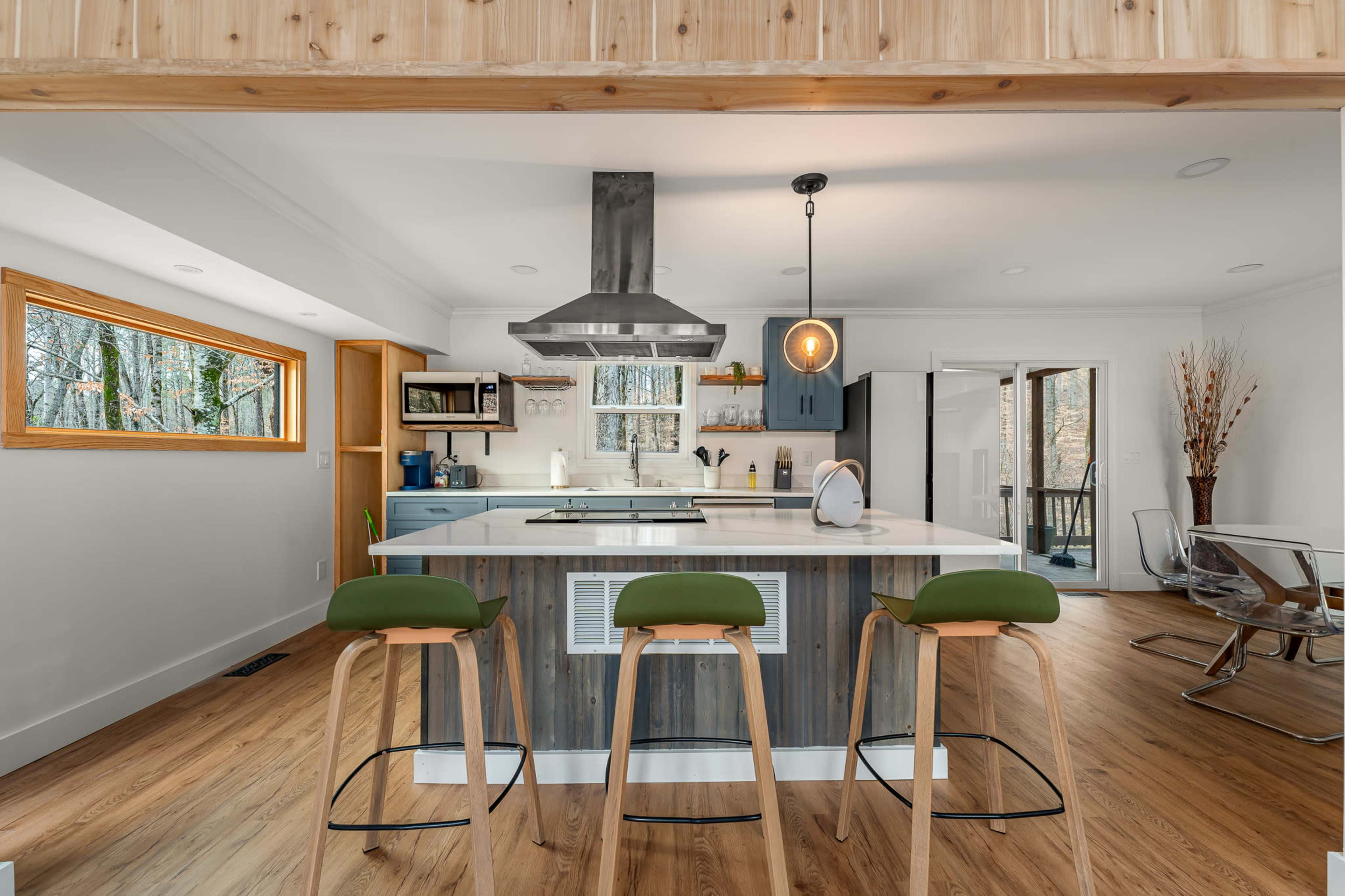 A modern kitchen features a central island with three stools, a stainless steel hood over a stovetop, and large windows showcasing a view of greenery outside.