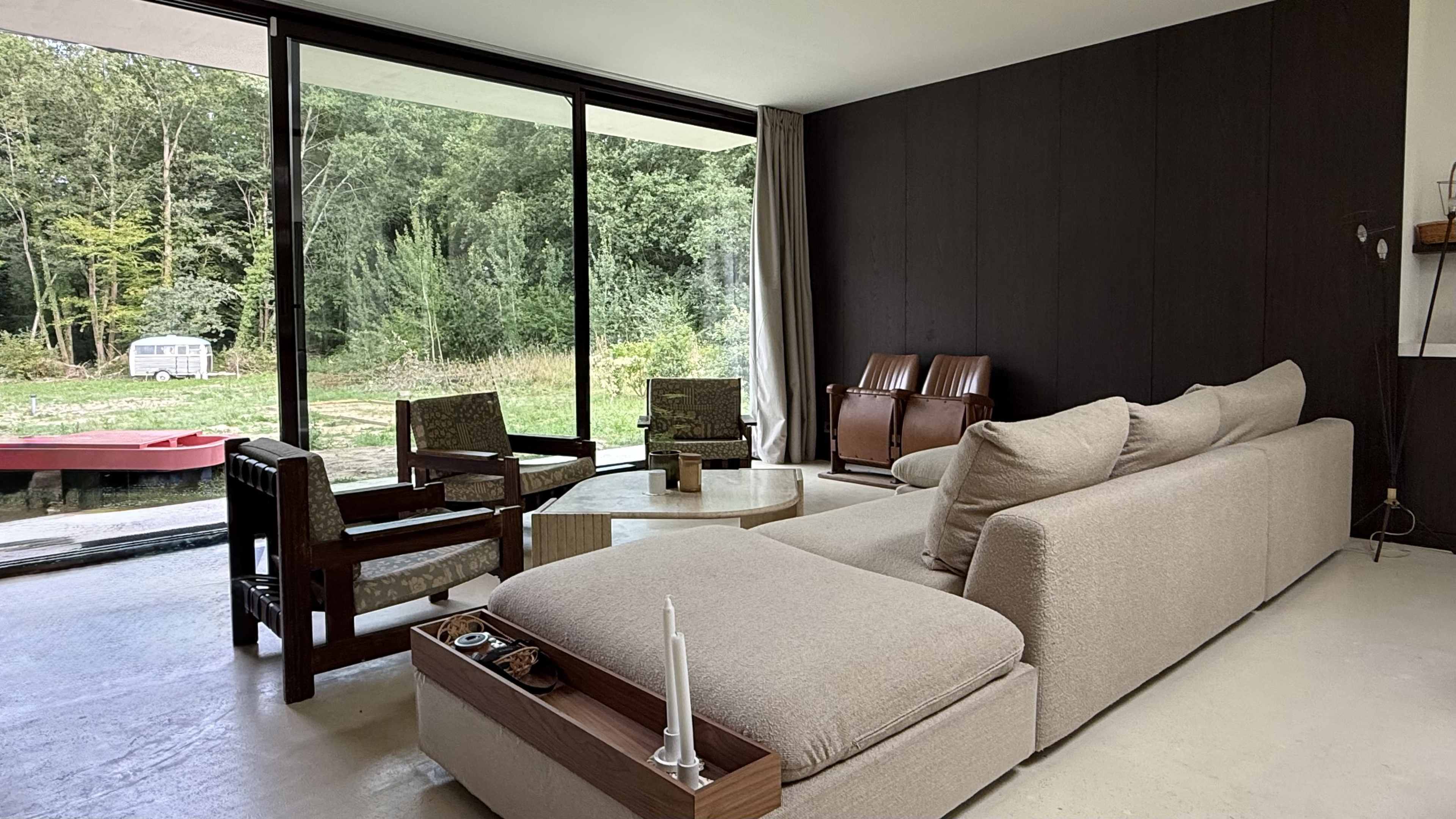The image shows a modern living room with a sectional sofa, two armchairs, a round table, and large glass windows looking out into a green forested area.