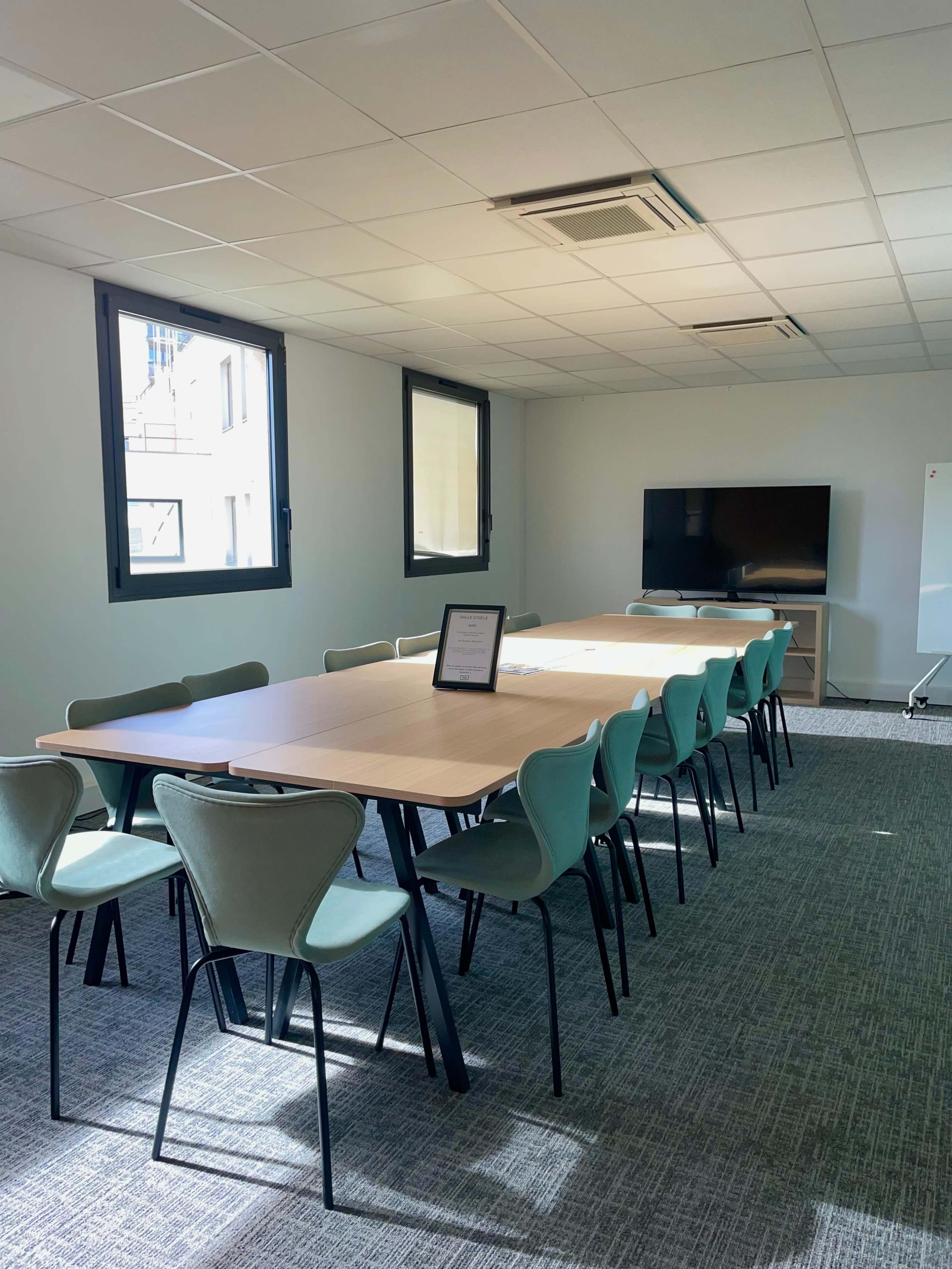 A long conference table with teal chairs is positioned in a well-lit meeting room featuring a large TV and a whiteboard.