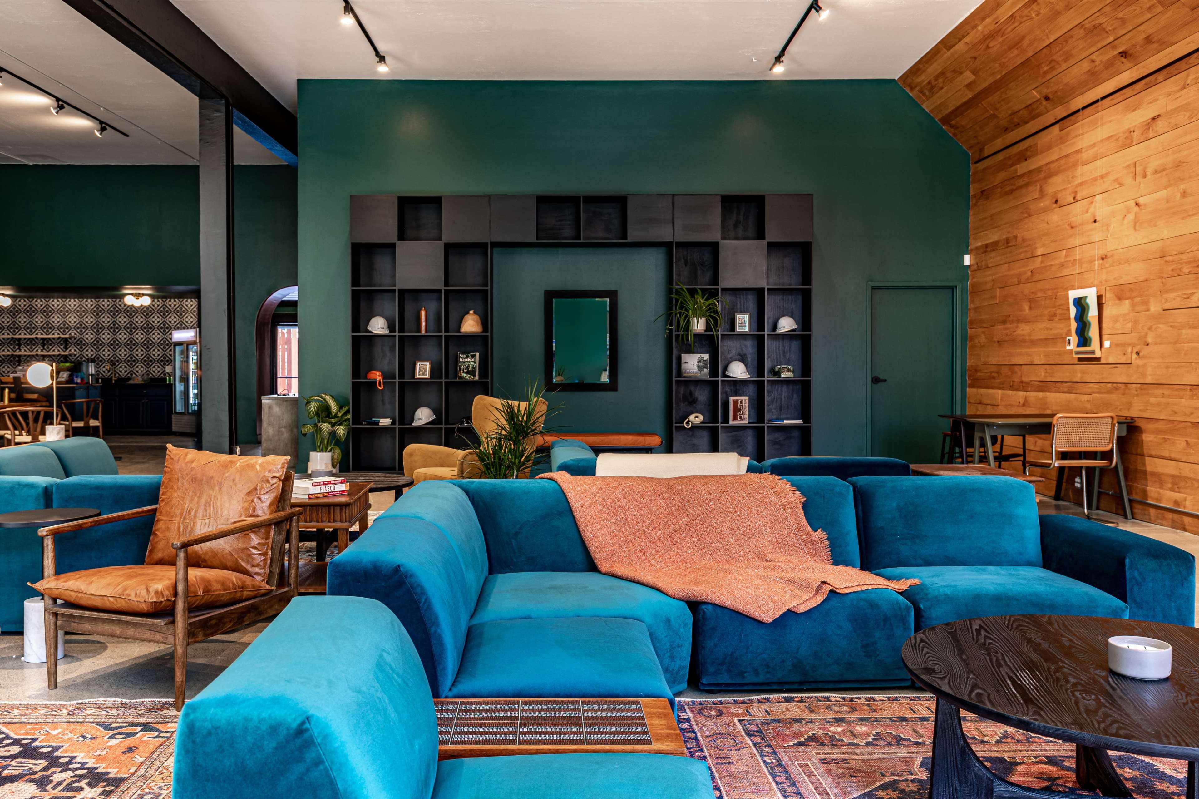 A spacious, stylish lounge area features a large teal sectional sofa, wooden accents, and decorative shelves against a deep green wall.