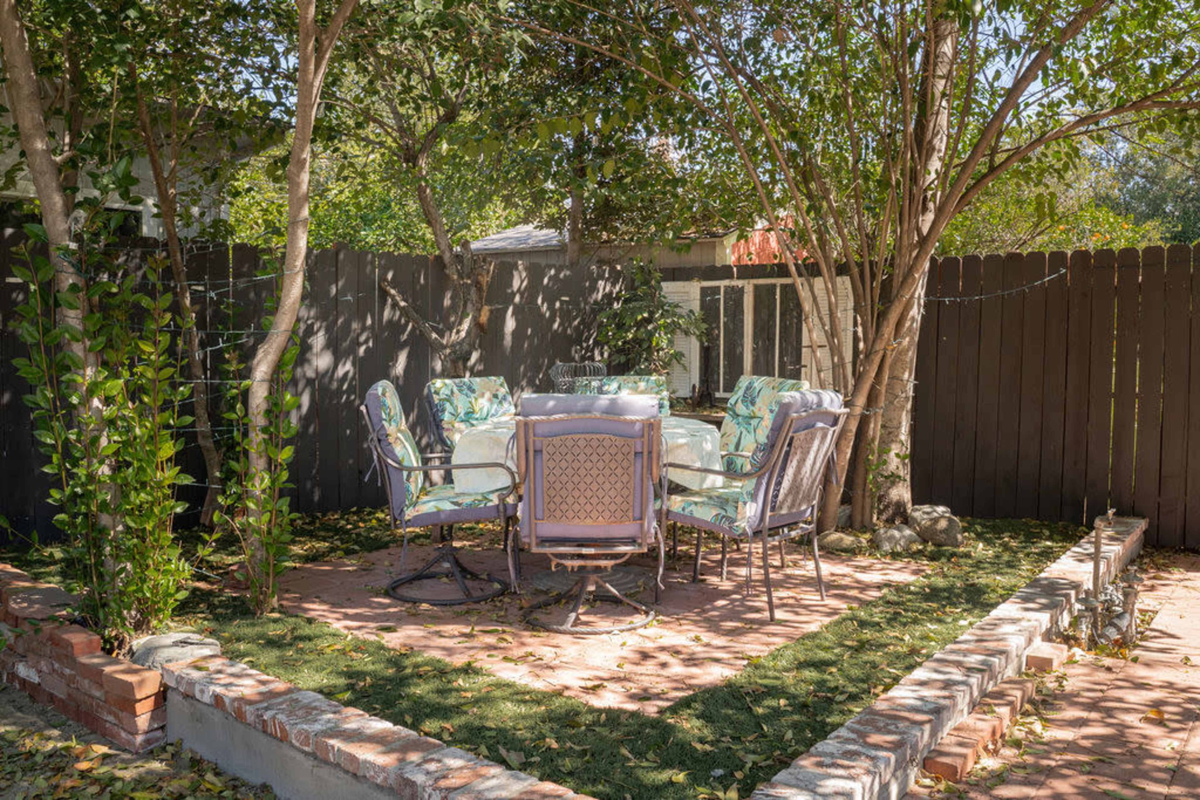 A circular patio area with a glass table surrounded by metal chairs is shaded by trees, enclosed by a wooden fence.