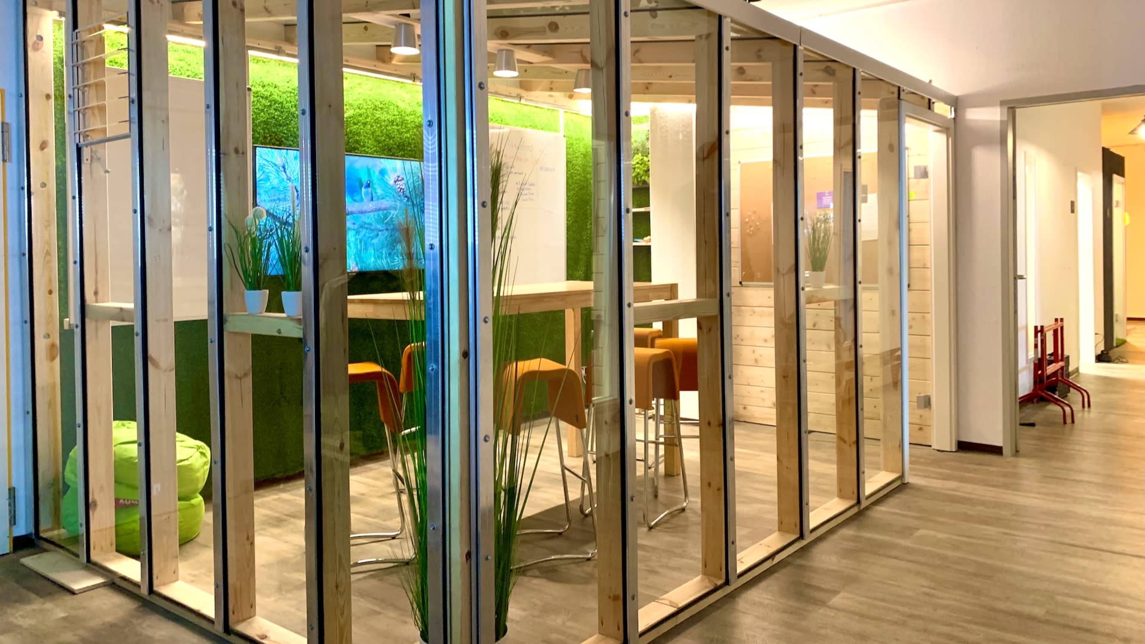 The image shows a modern office space featuring wooden-framed enclosures, green wall accents, and a small seating area with orange chairs.